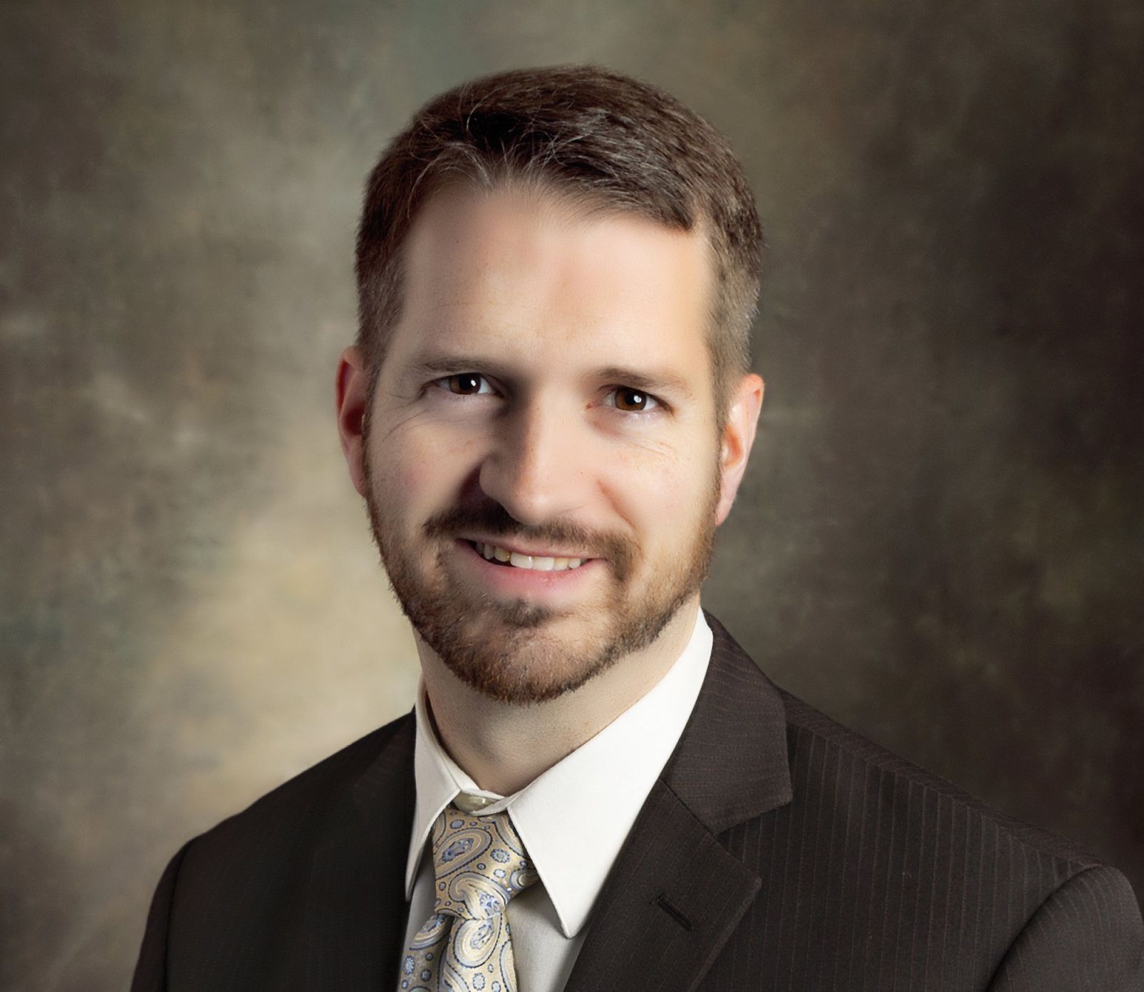 A person with a short beard smiles while wearing a dark suit, white collared shirt, and a patterned tie against a backdrop.