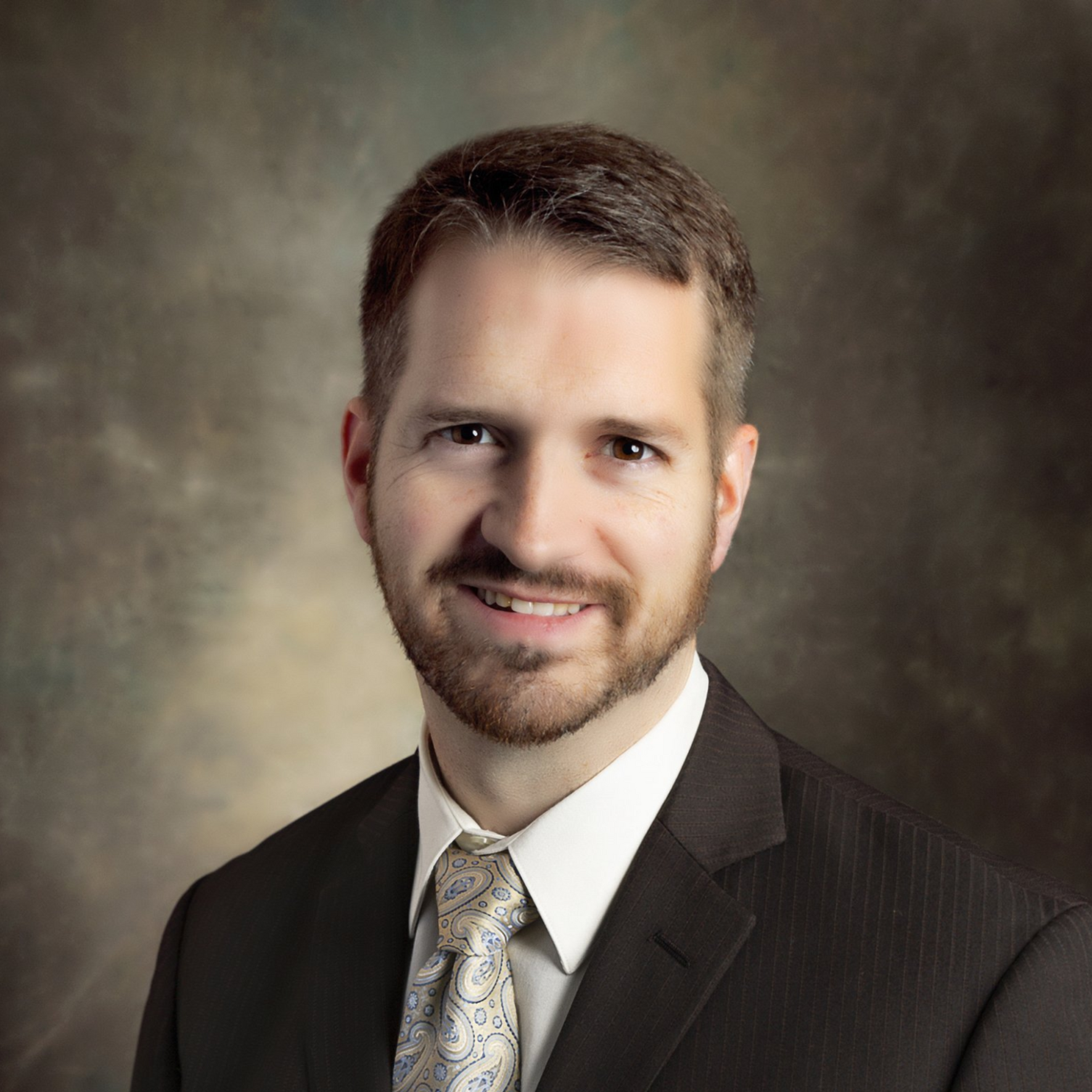 A person with short brown hair and a beard, wearing a dark suit, white shirt, and patterned tie against a mottled backdrop.