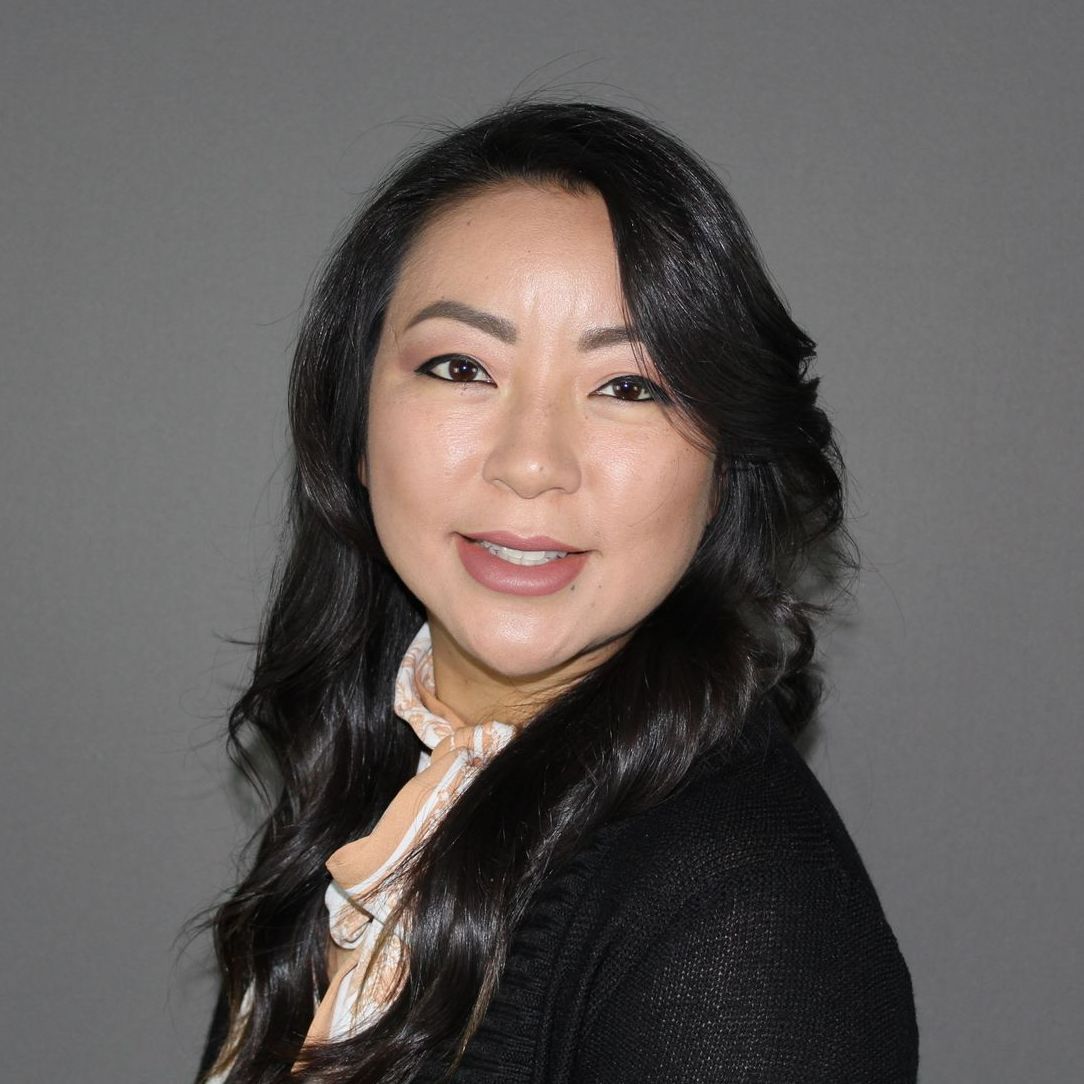A portrait of a person with long, wavy dark hair, wearing a patterned scarf and black top against a gray background.