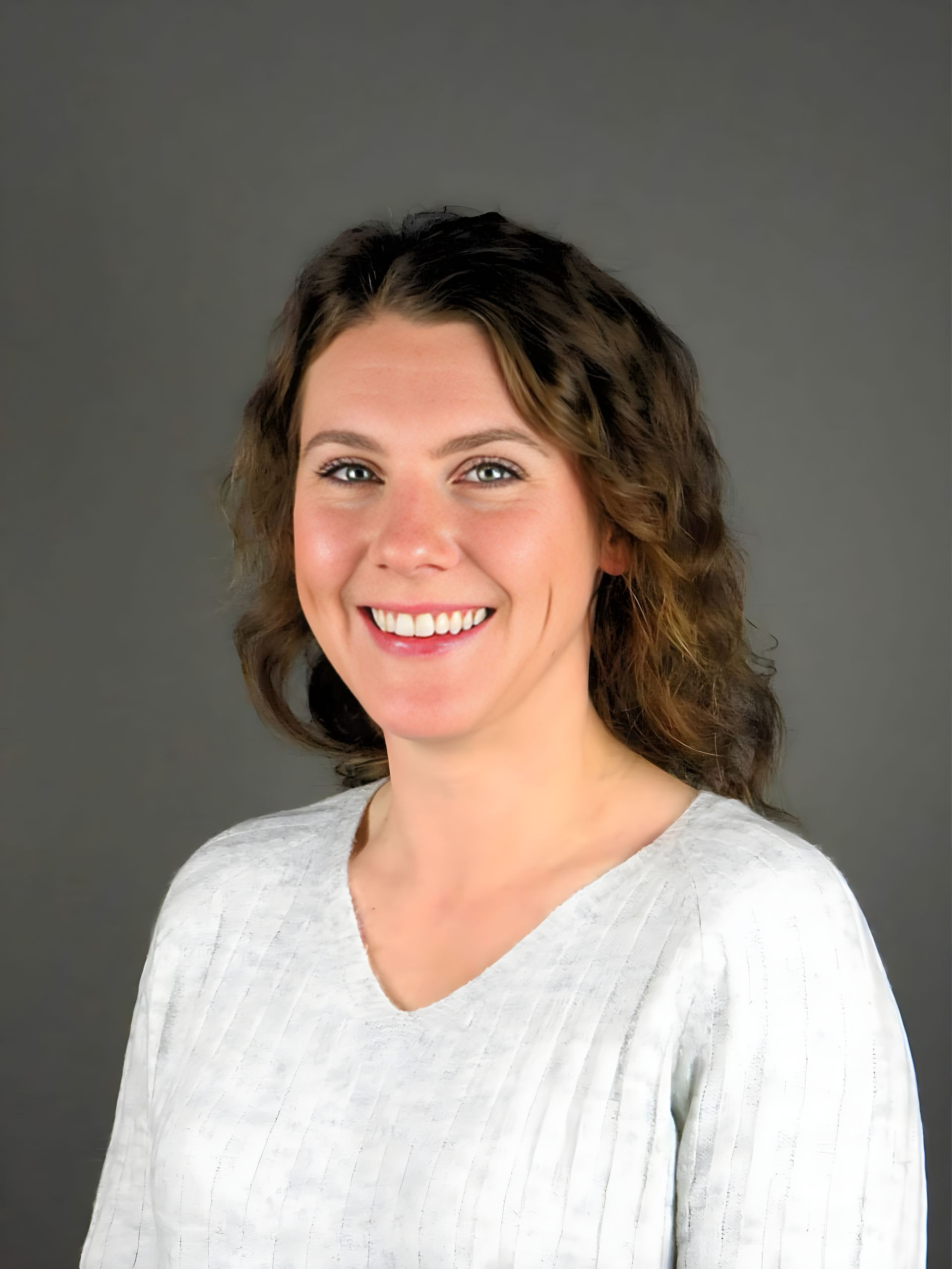 A smiling person with wavy brown hair wearing a textured light-colored V-neck sweater against a plain gray background.
