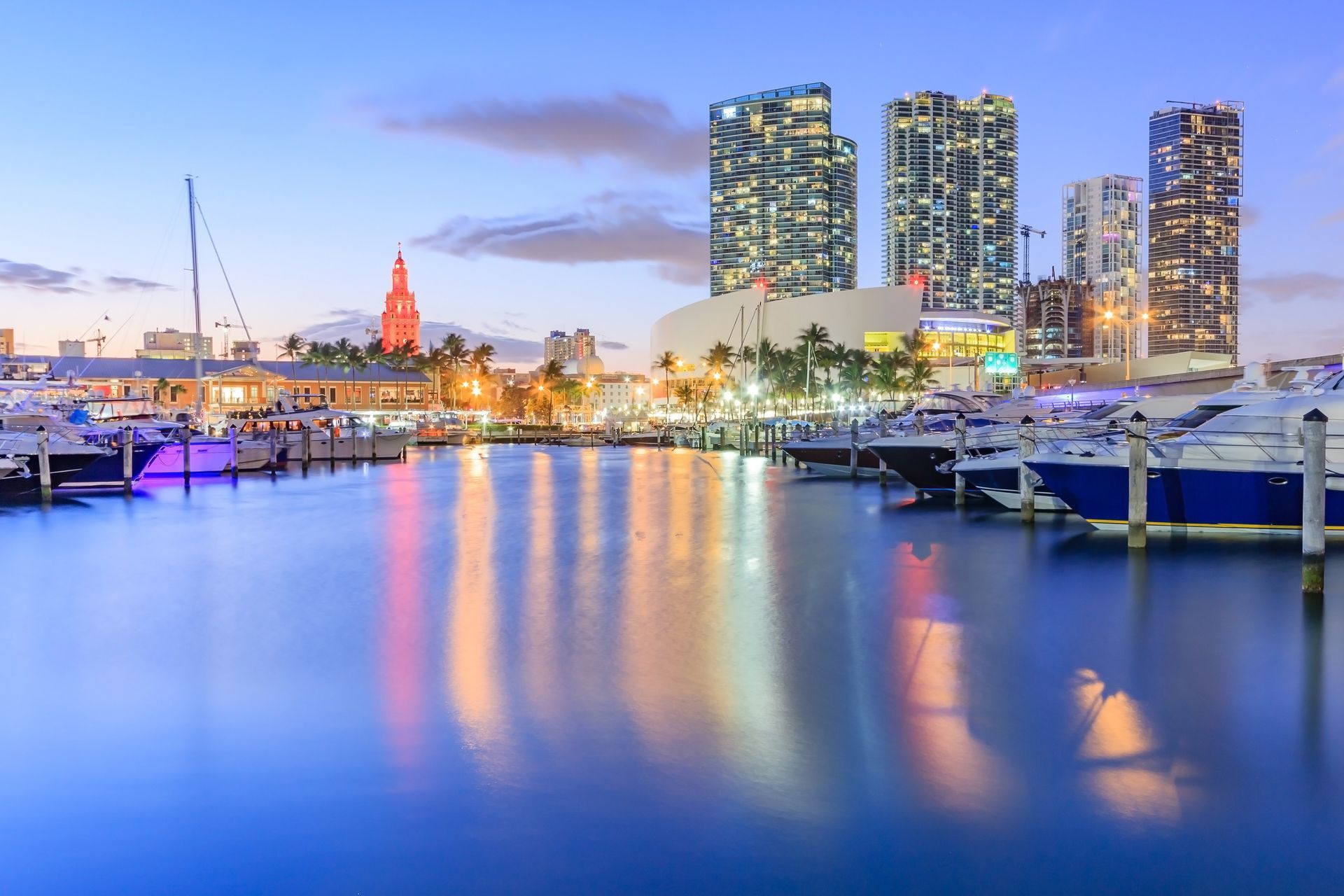 Nighttime waterfront city skyline with lit buildings, marina boats, and colorful reflections on calm water