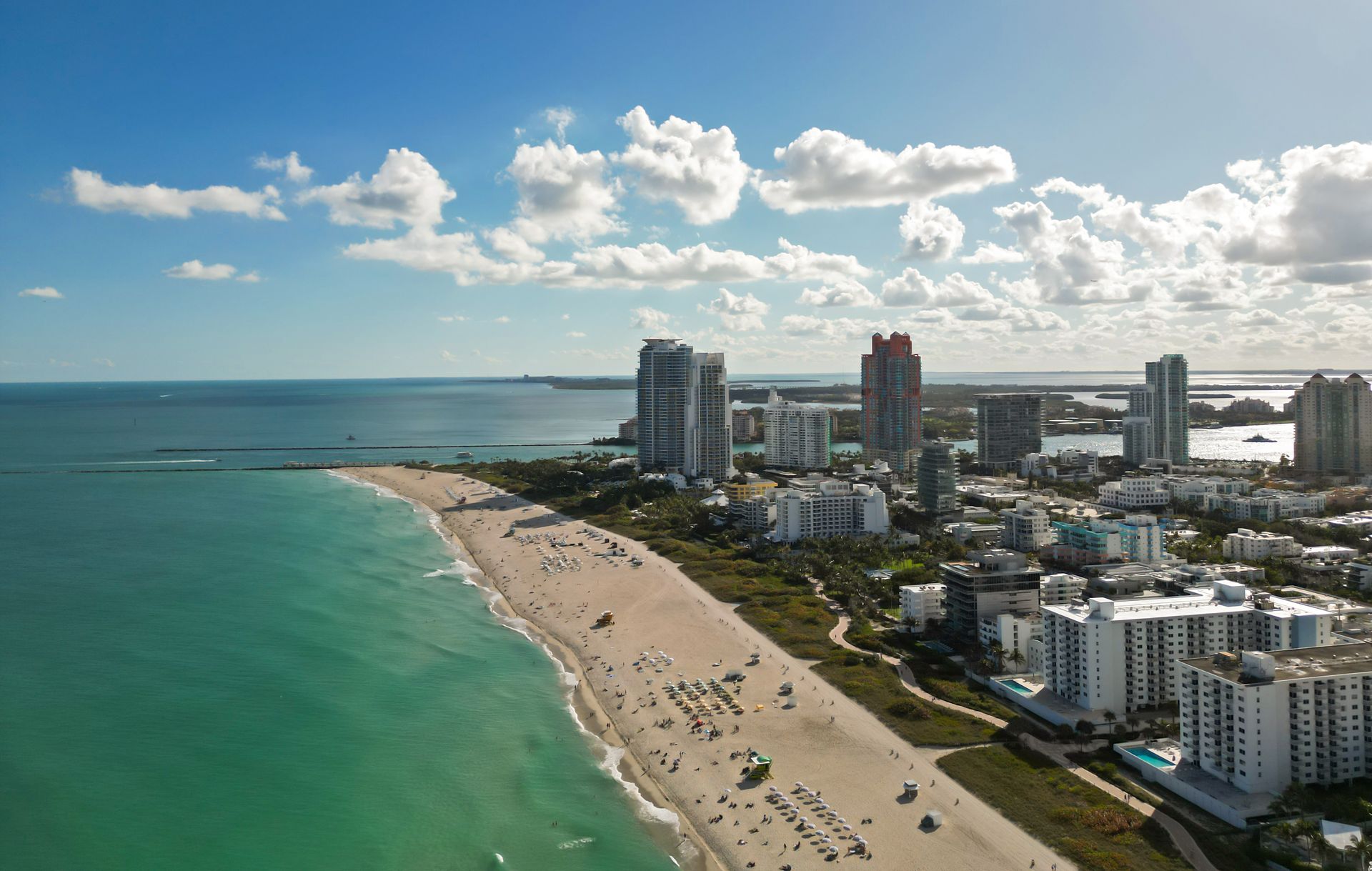 Aerial view of a sandy beach, turquoise ocean, and high-rise buildings under a blue sky with clouds
