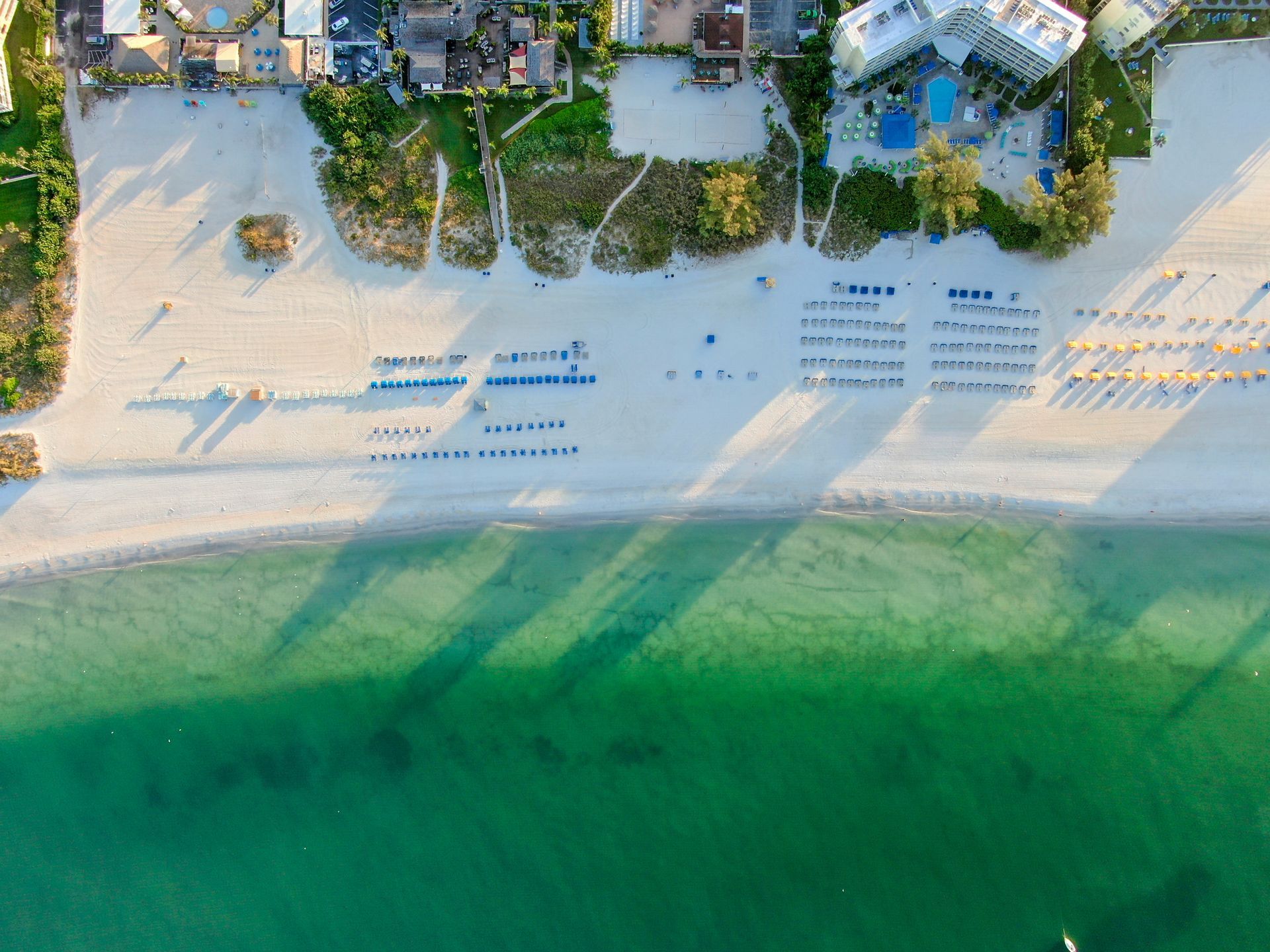 Aerial view of a sandy beach with rows of lounge chairs, green water, and resort buildings at the top.