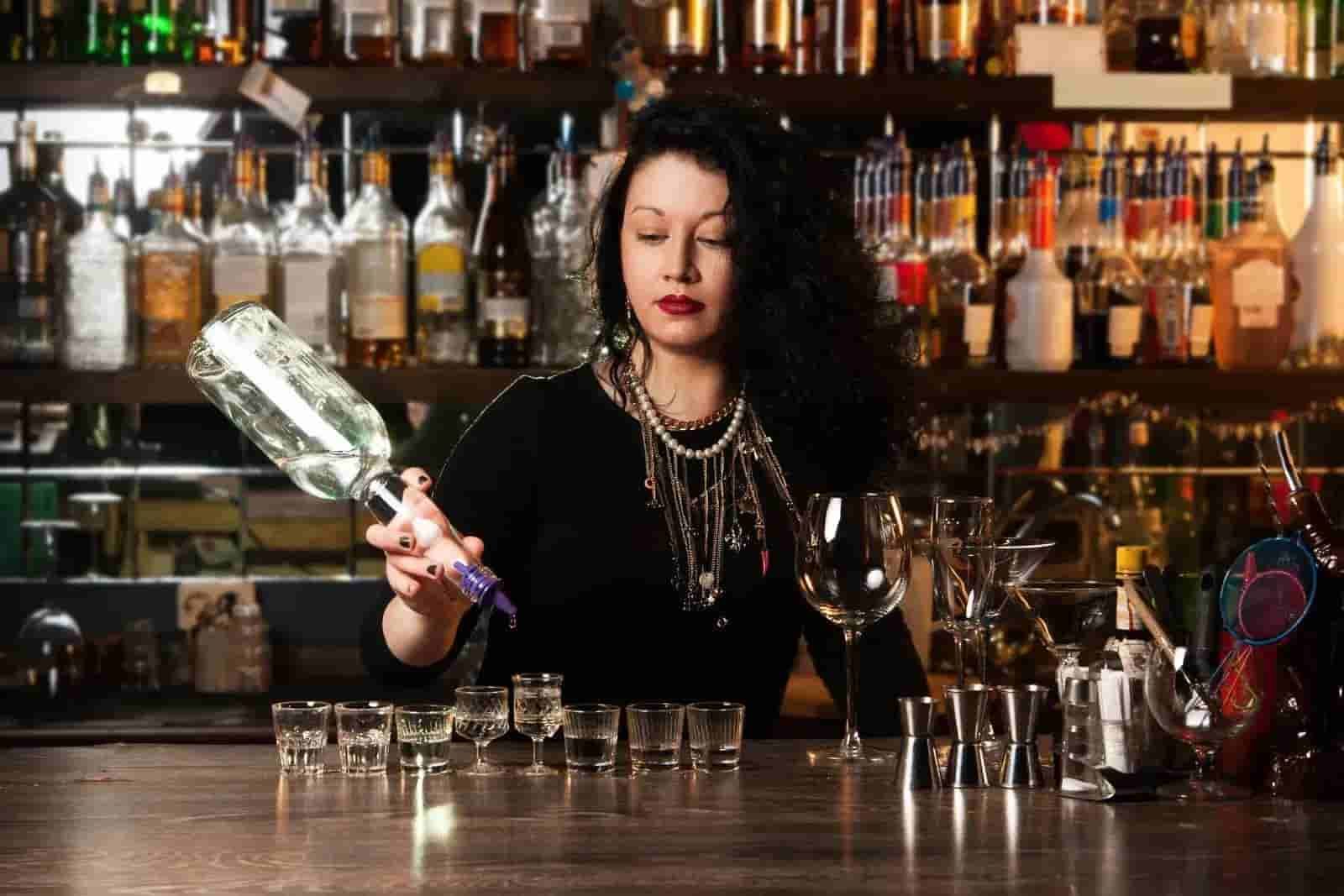 Bartender in black dress mixing a cocktail behind a bar lined with bottles and glassware.