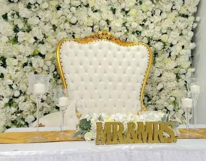 White tufted chair with gold trim before a white floral wall, with gold “MR & MRS”