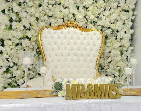 White tufted chair with gold trim before a white floral wall, with gold “MR & MRS”