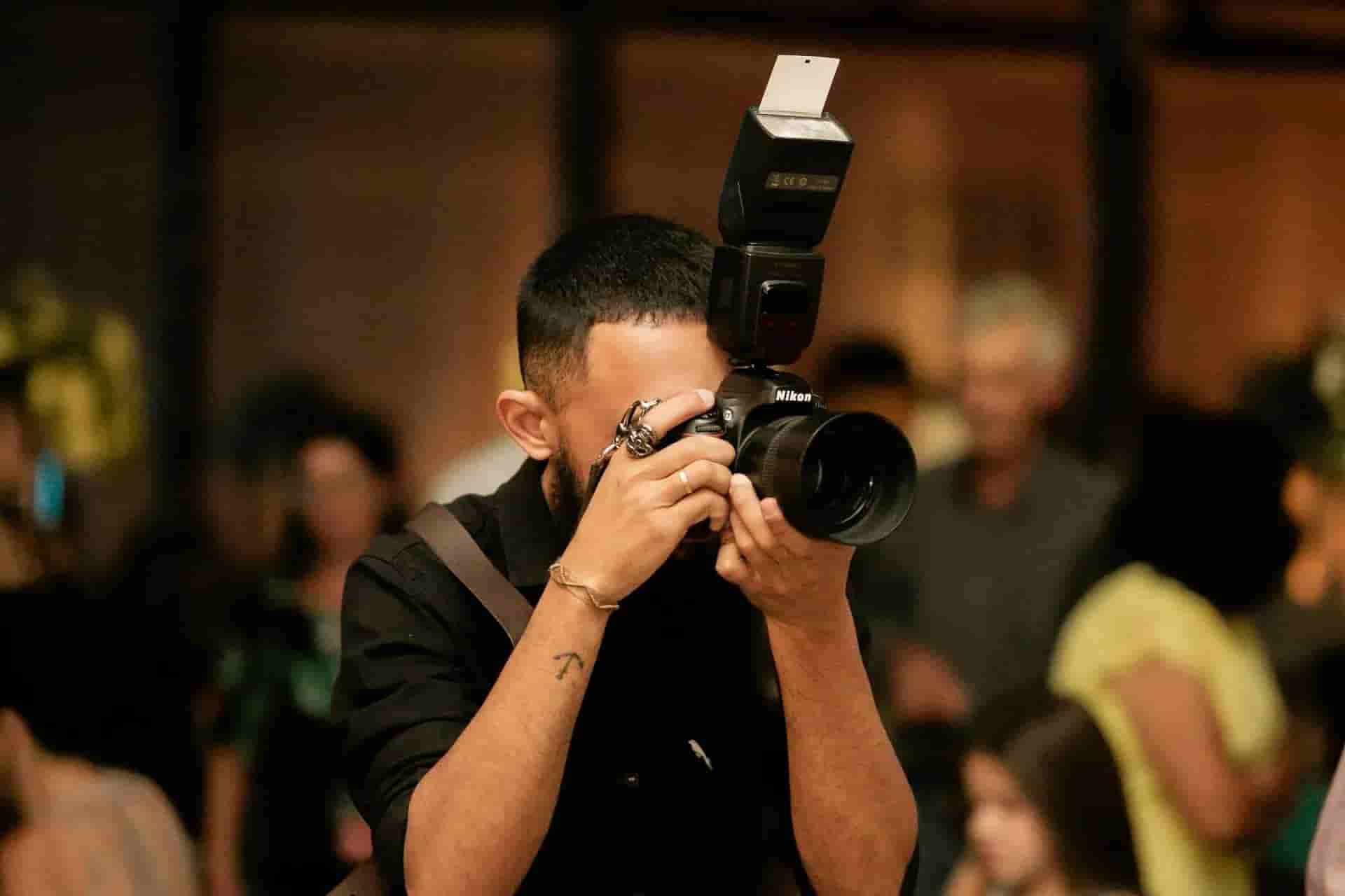 Photographer in black shirt aiming a camera with flash at an event, blurred people in background