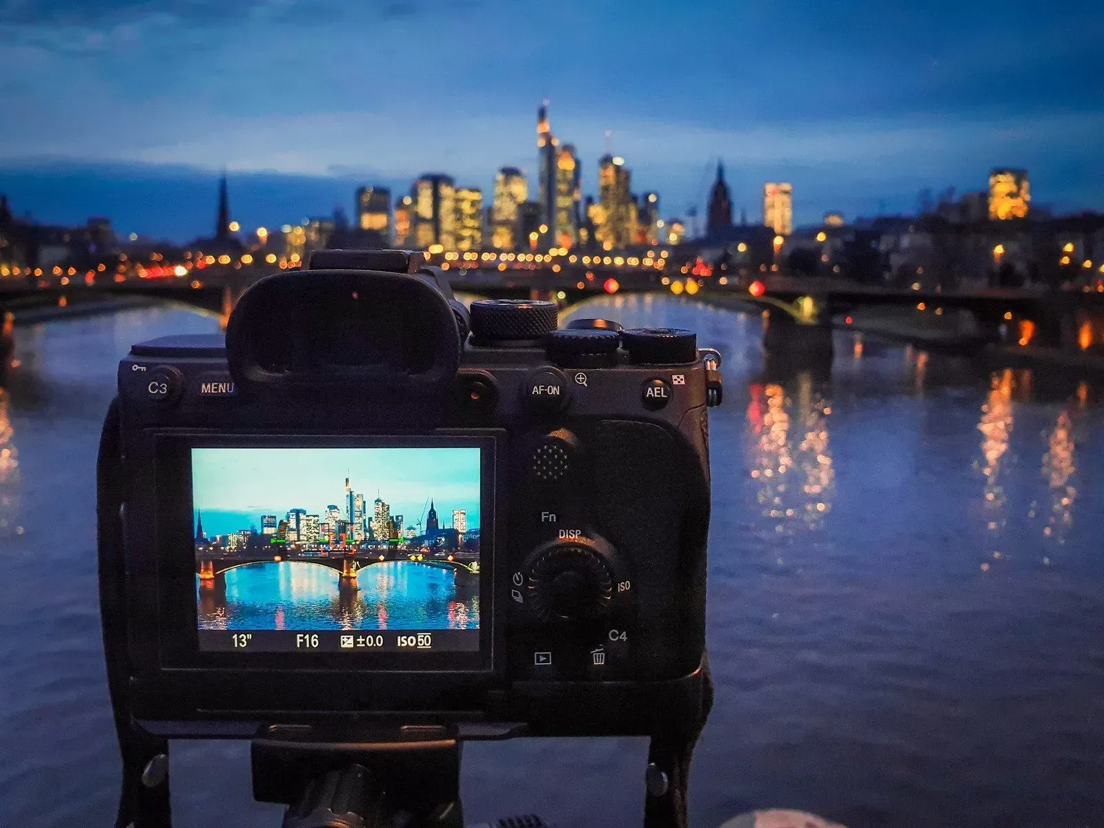 Camera framing a lit city skyline and river at dusk, reflected in the water.