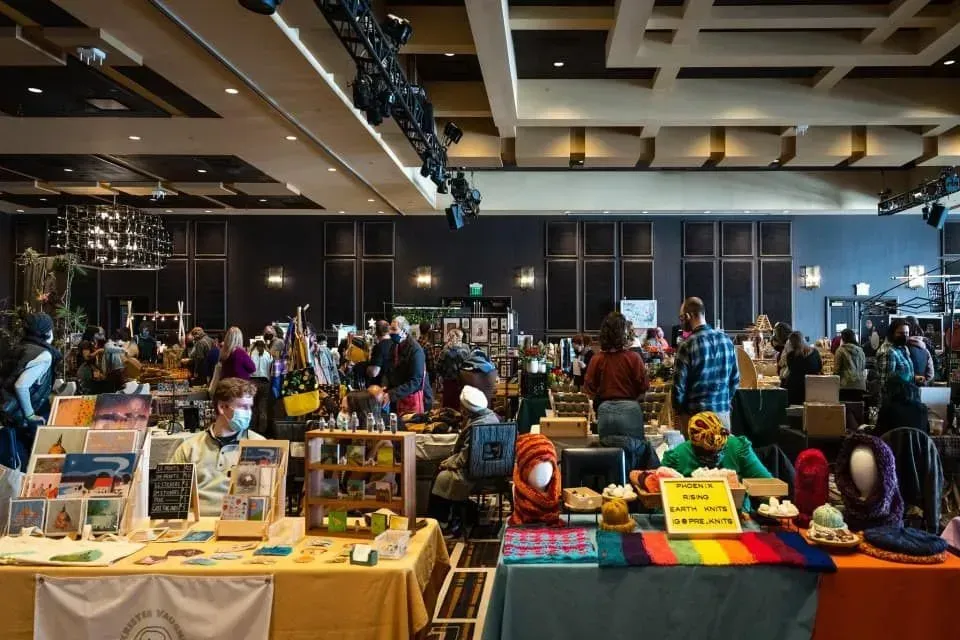 Indoor craft fair with crowded vendor tables displaying handmade goods and colorful items