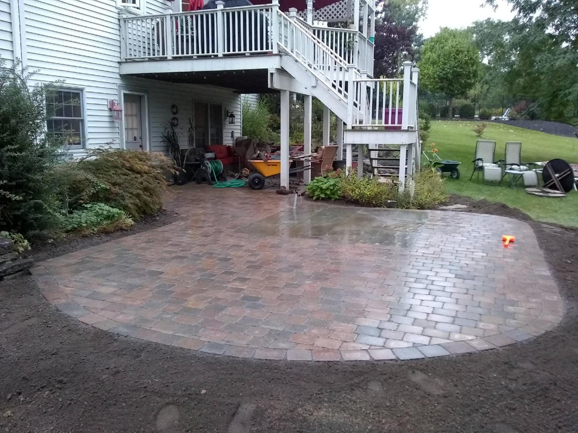 Brick patio below a two-story deck, bordered by soil and plants.
