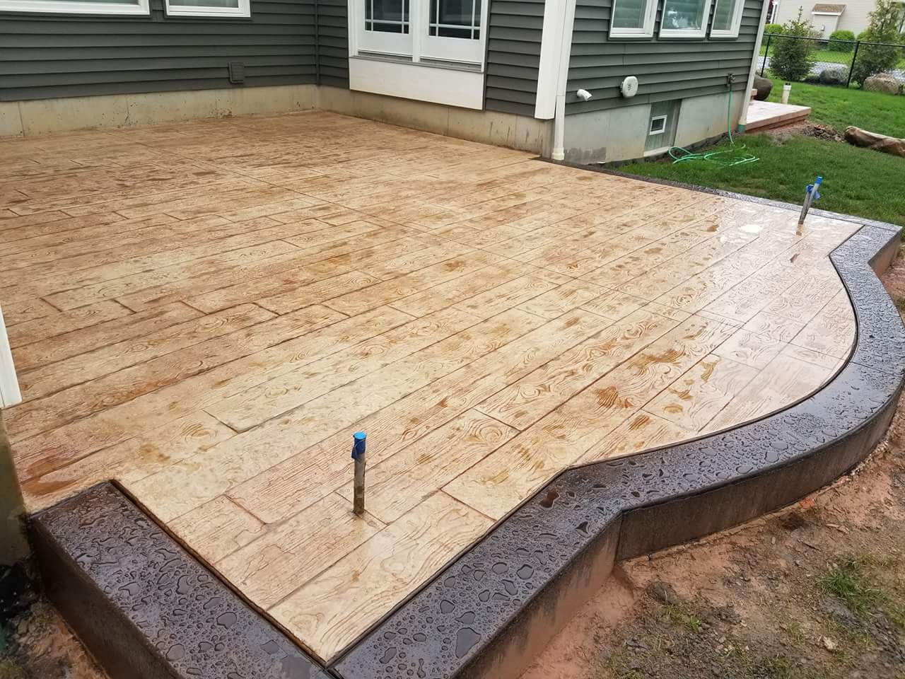 Concrete patio with a curved border, stamped to resemble wood planks, next to a house.
