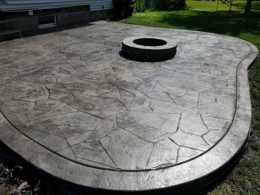 Stone-textured concrete patio with a fire pit in the center. The patio borders a grassy yard, partially in sunlight.