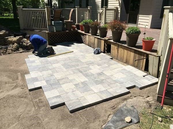 A worker laying gray paving stones for a patio; a deck and potted plants are in the background.