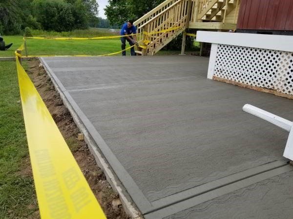 Freshly poured concrete patio with a worker in the background near a house. Yellow caution tape surrounds the perimeter.