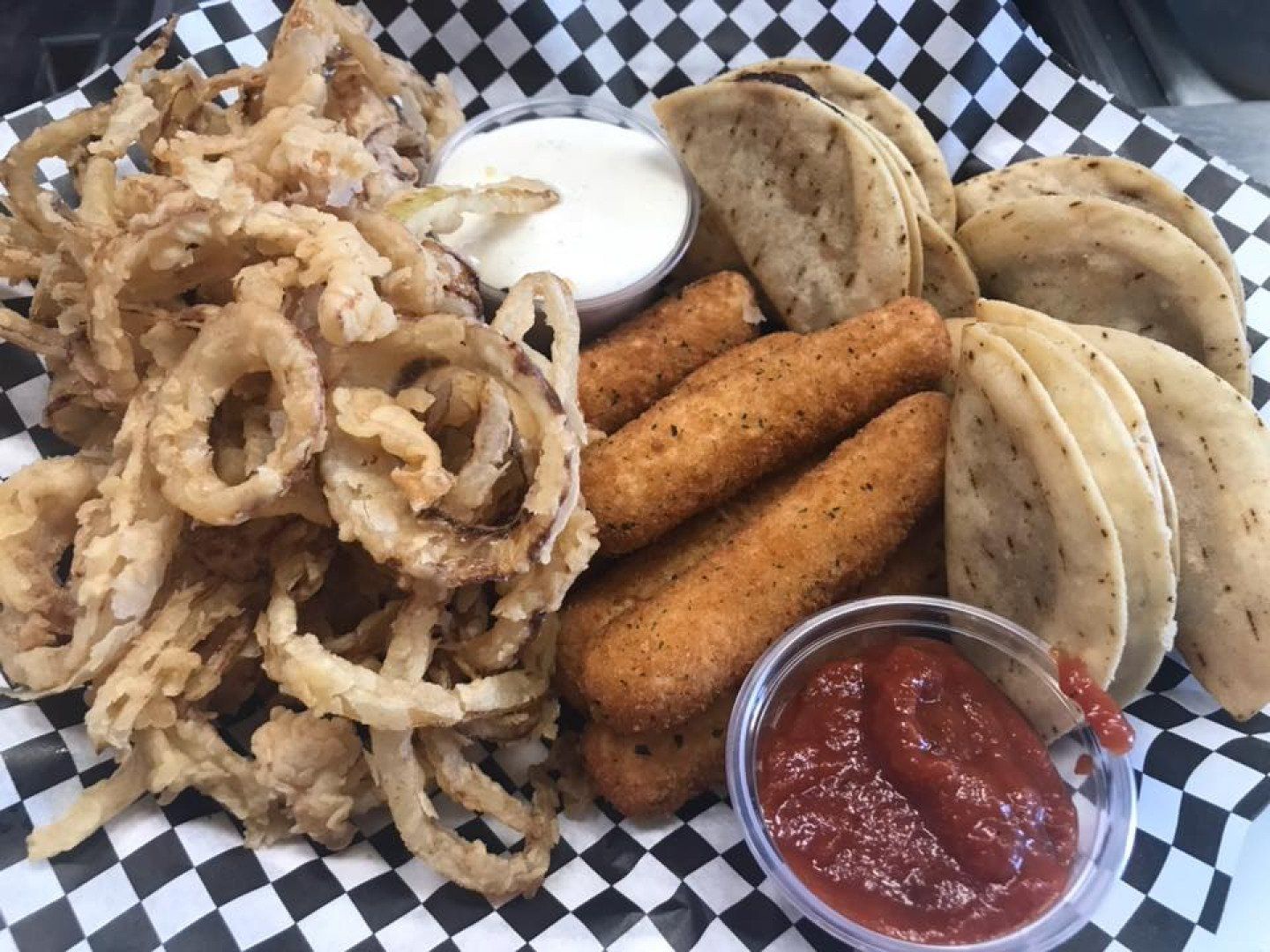 A plate of food with onion rings , mozzarella sticks , and tacos on a checkered paper.