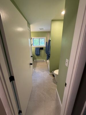 Narrow hallway with open bathroom, pale green walls, and a window at the end.