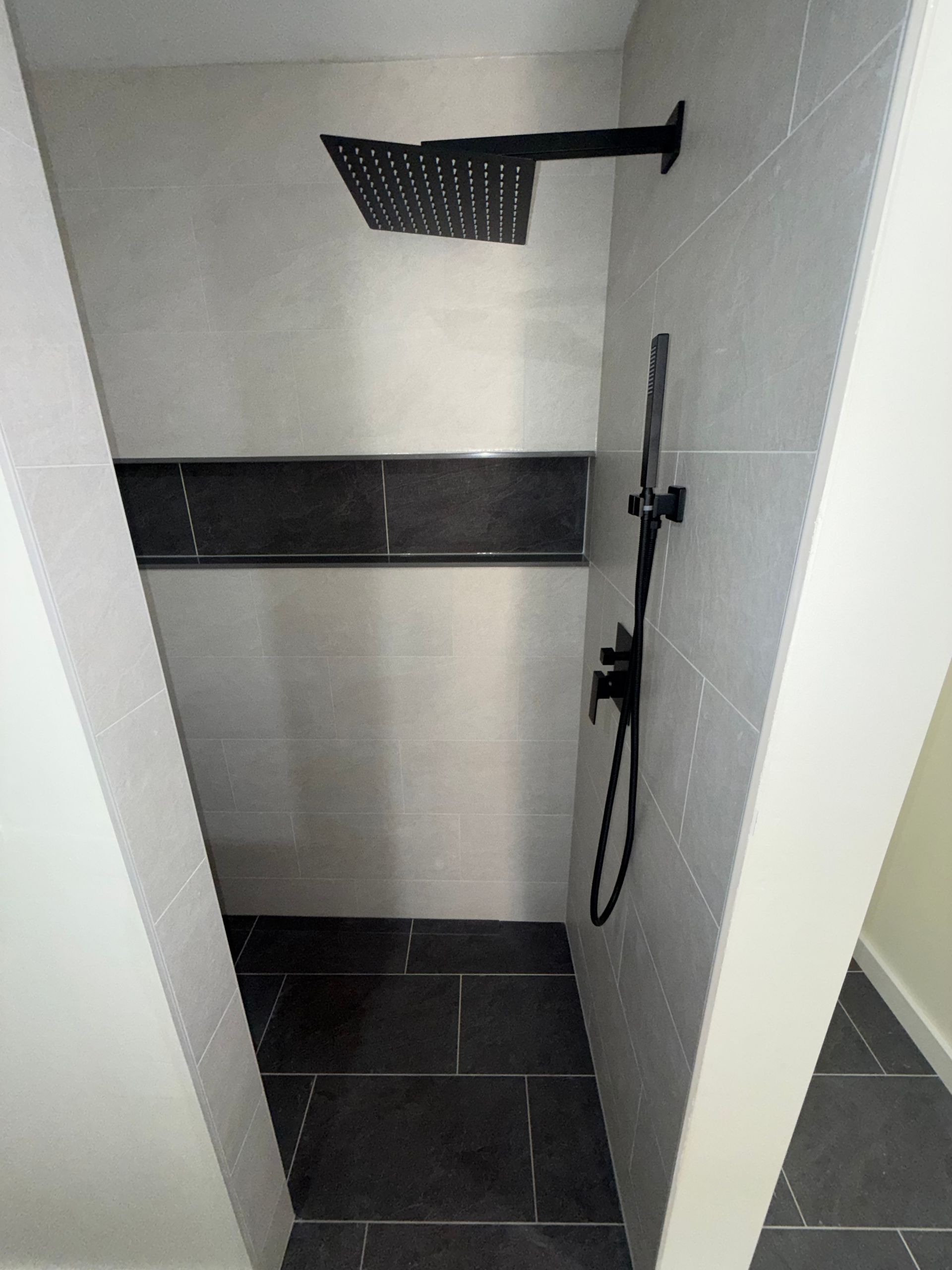Small tiled shower stall with black floor, wall shelf, overhead showerhead, and handheld shower on the right.