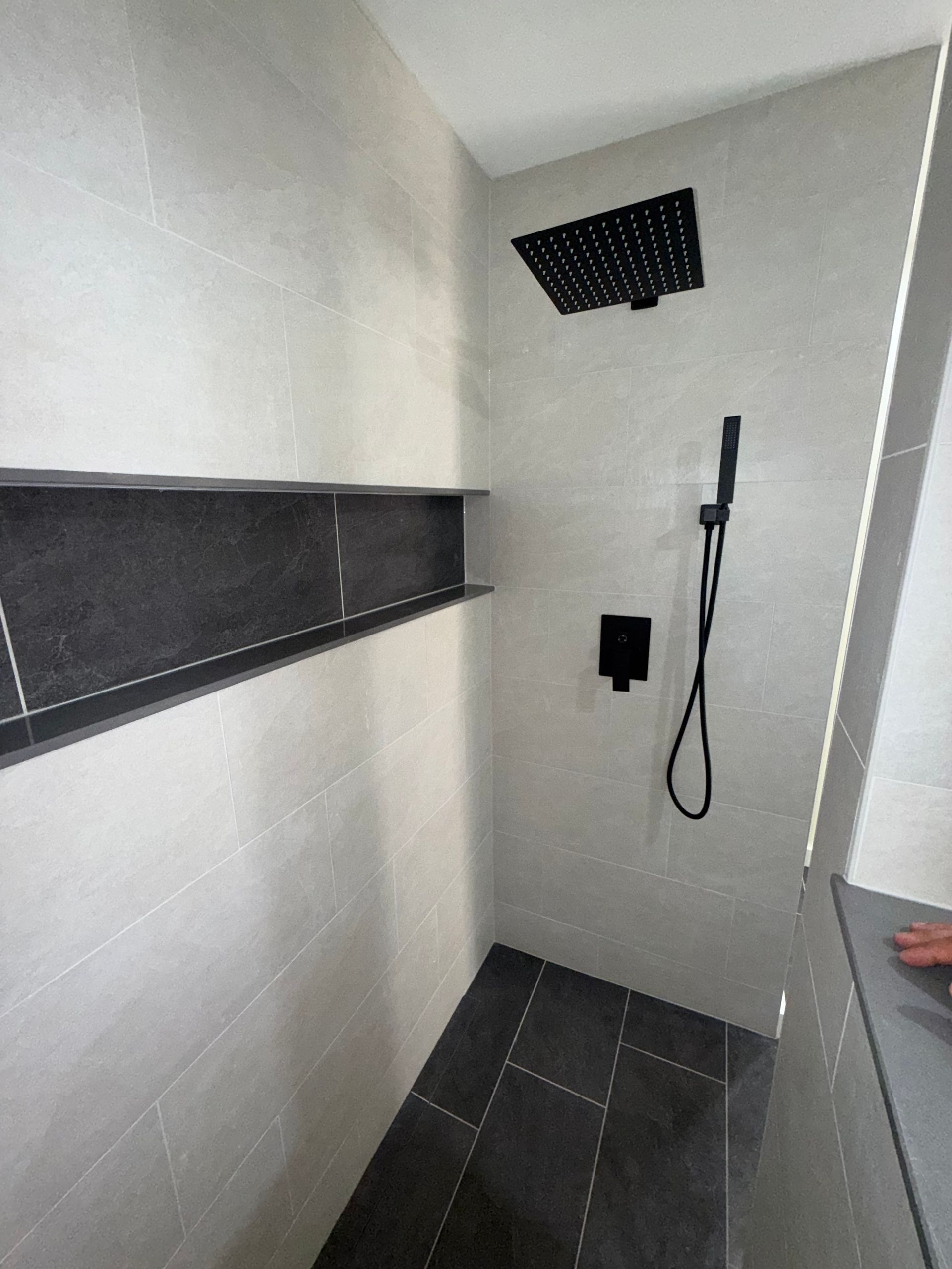 Small tiled shower stall with a black showerhead, shelf, and floor drain.