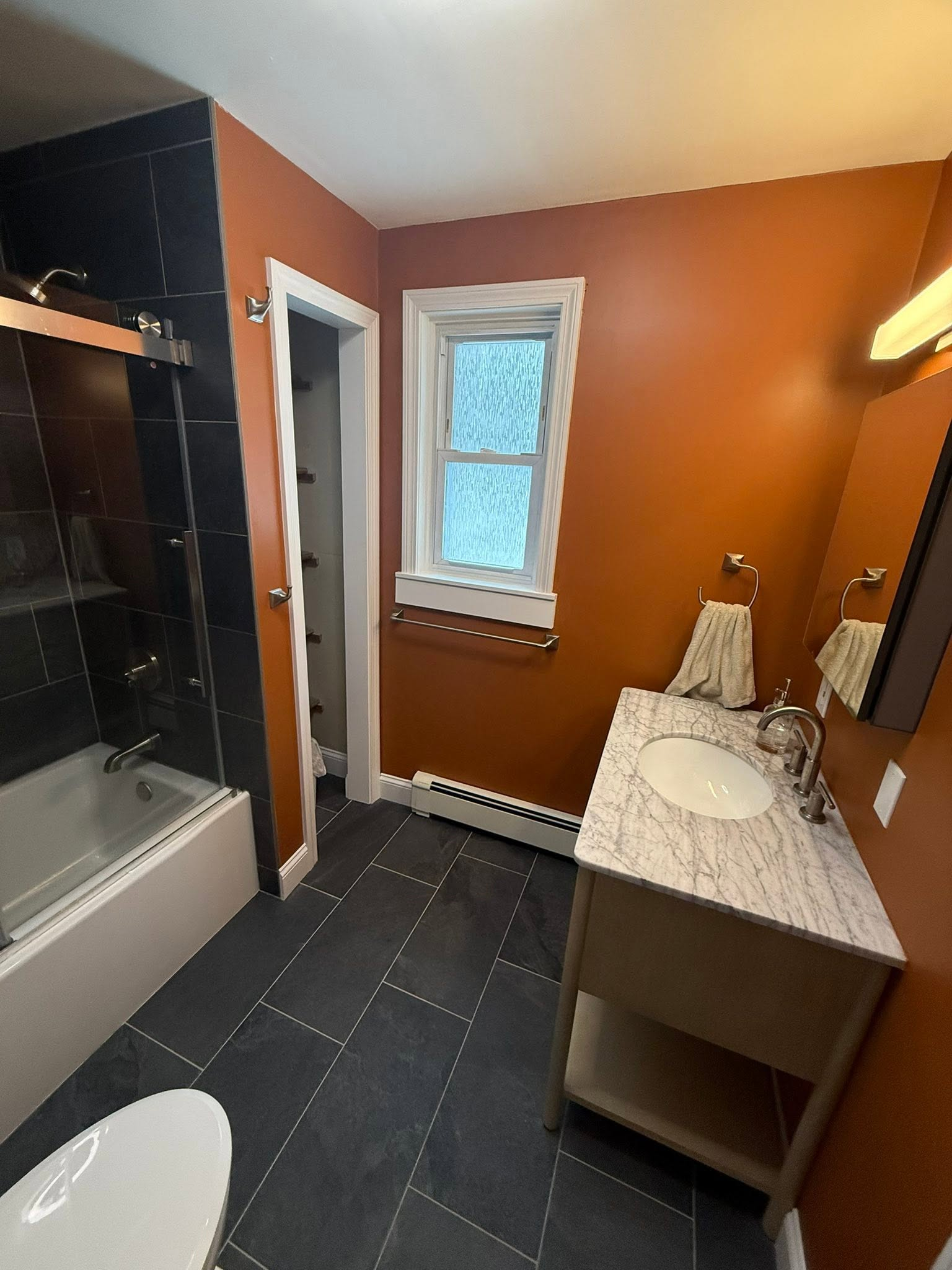 Small bathroom with orange walls, black tiled floor, shower, sink vanity, and window.
