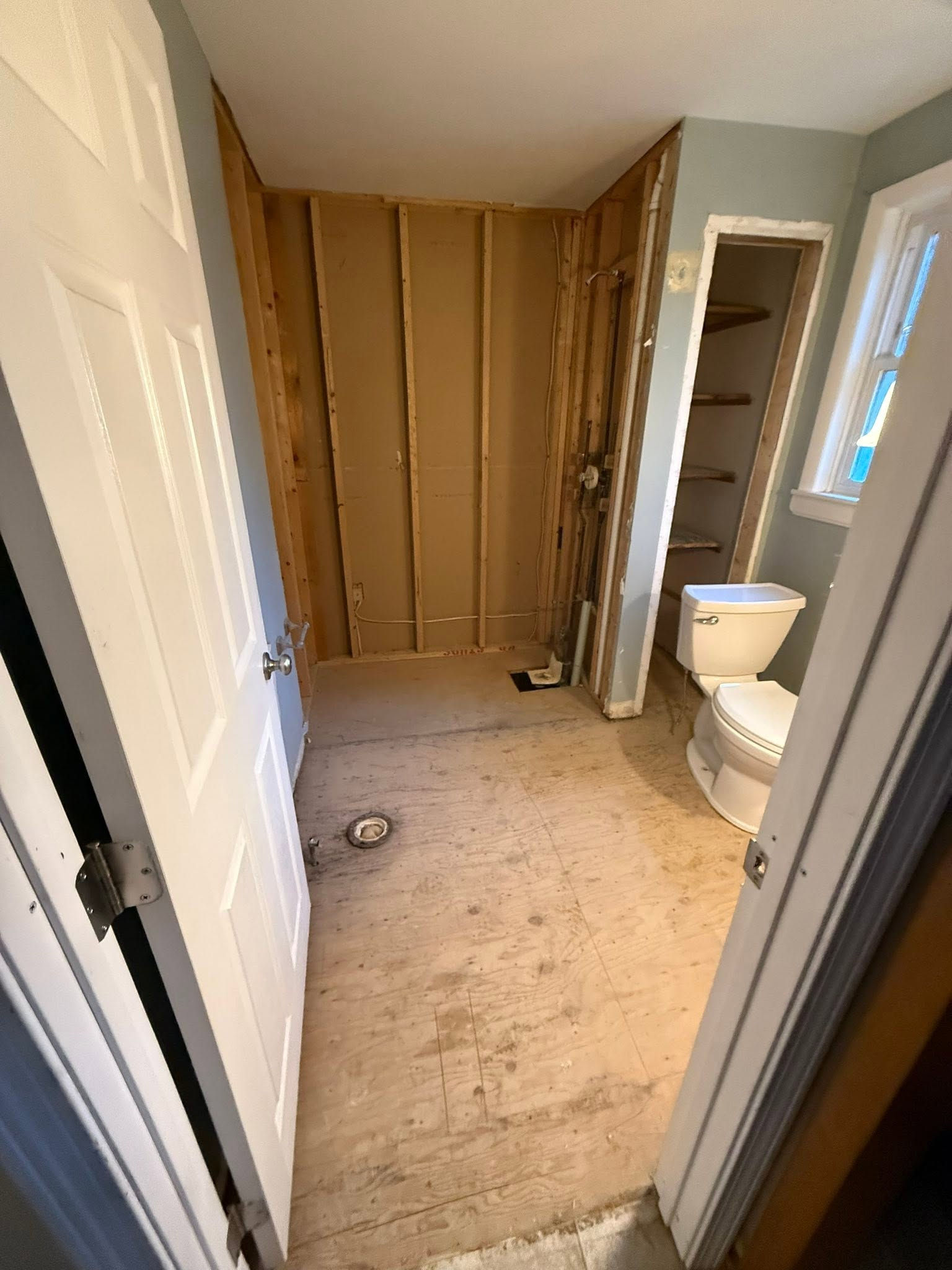 Bathroom under renovation with exposed studs, bare subfloor, toilet, and built-in shelving by a window.