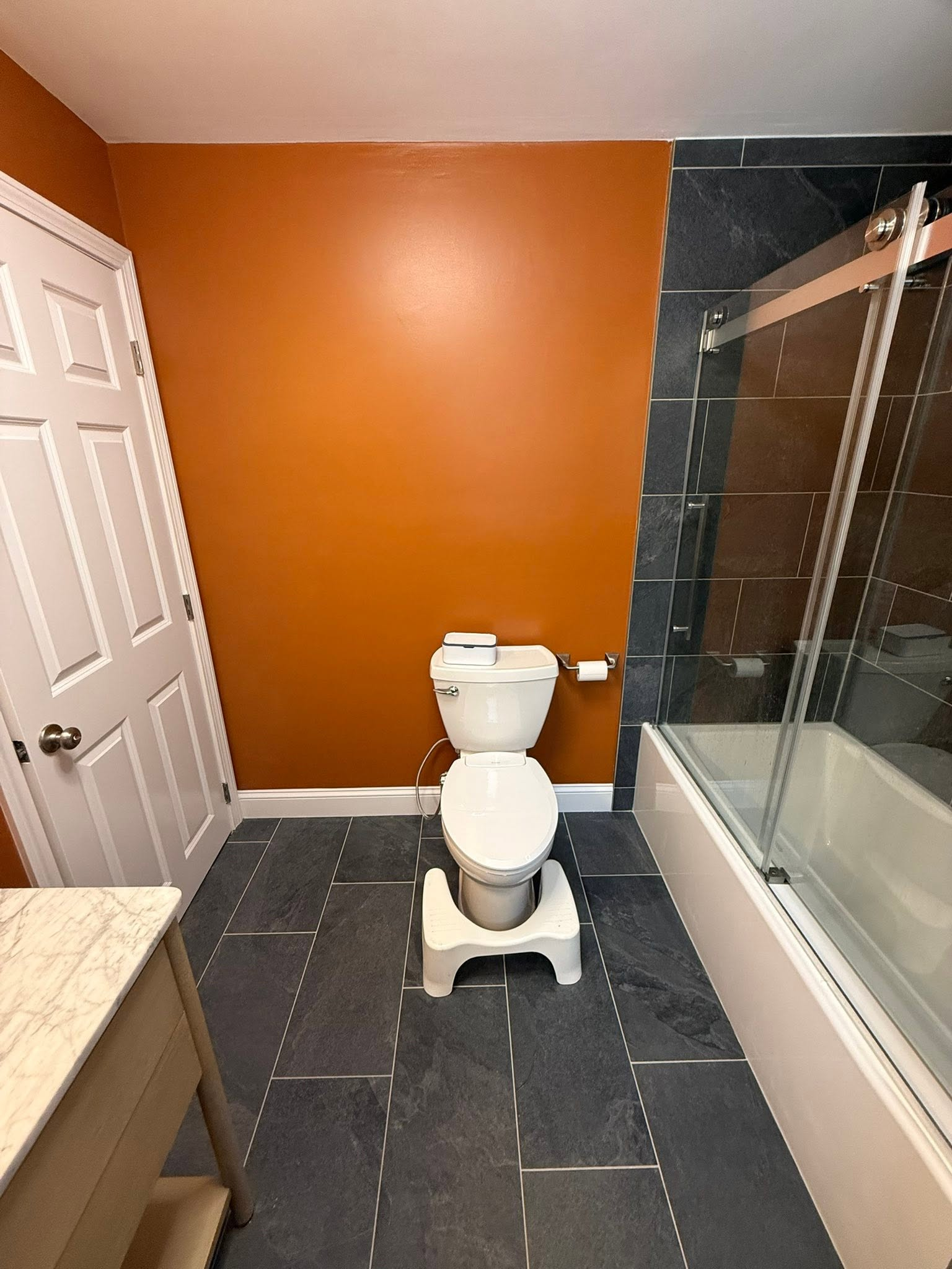 Small bathroom with orange wall, white toilet, and glass shower beside a bathtub.