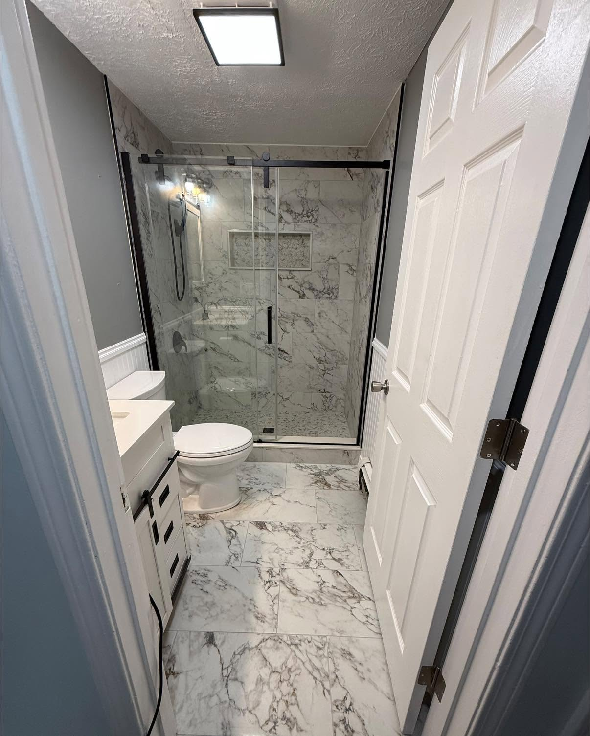 Modern bathroom with marble tile, glass shower, toilet, and white vanity, viewed through an open door