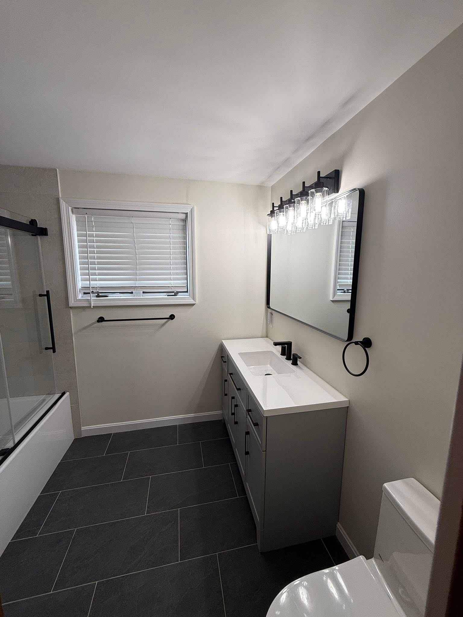 Small bathroom with gray vanity, mirror lights, window blinds, and white walls.