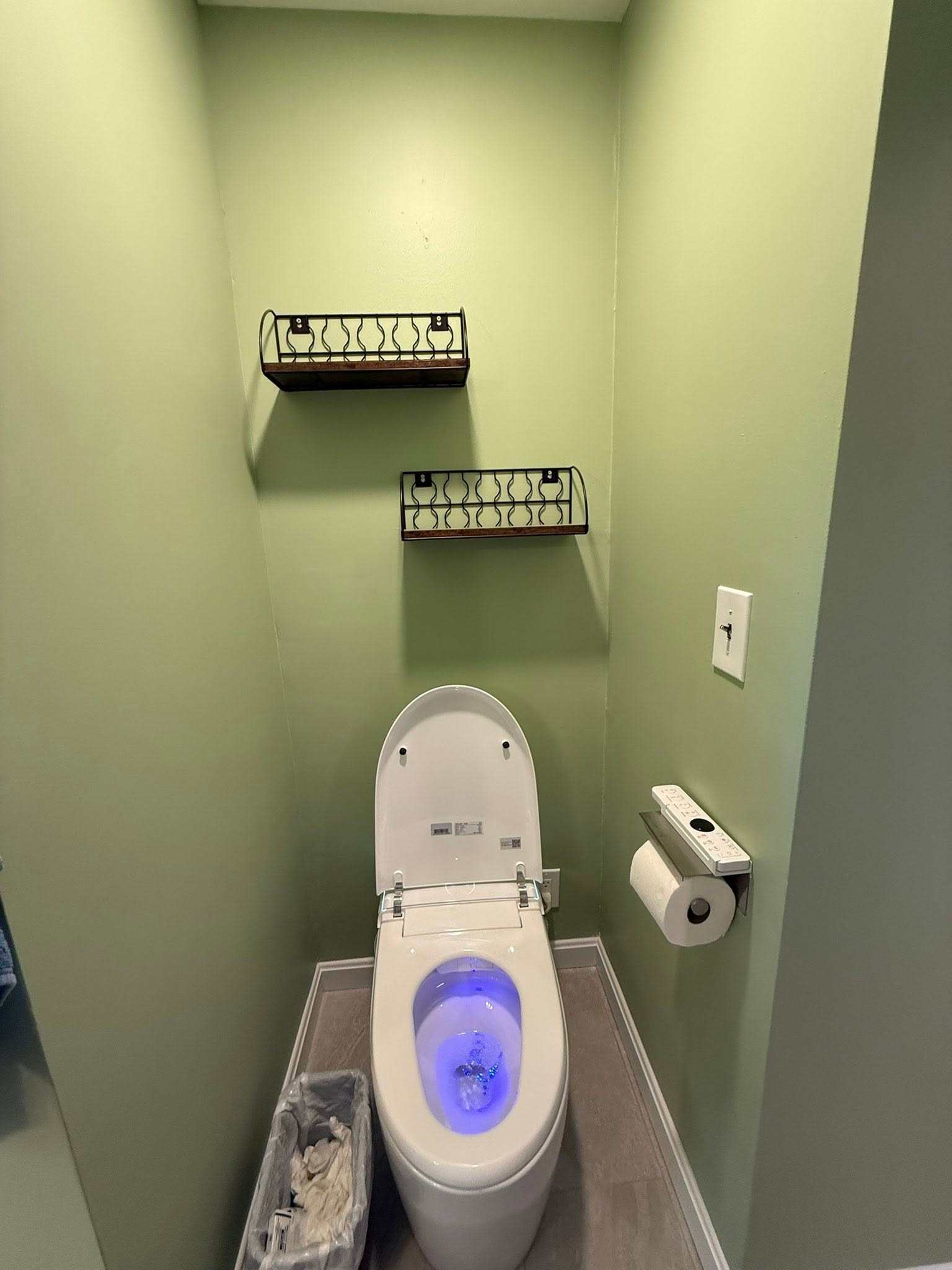 Small restroom with a toilet, glowing blue seat light, toilet paper holder, and wall shelves on green walls