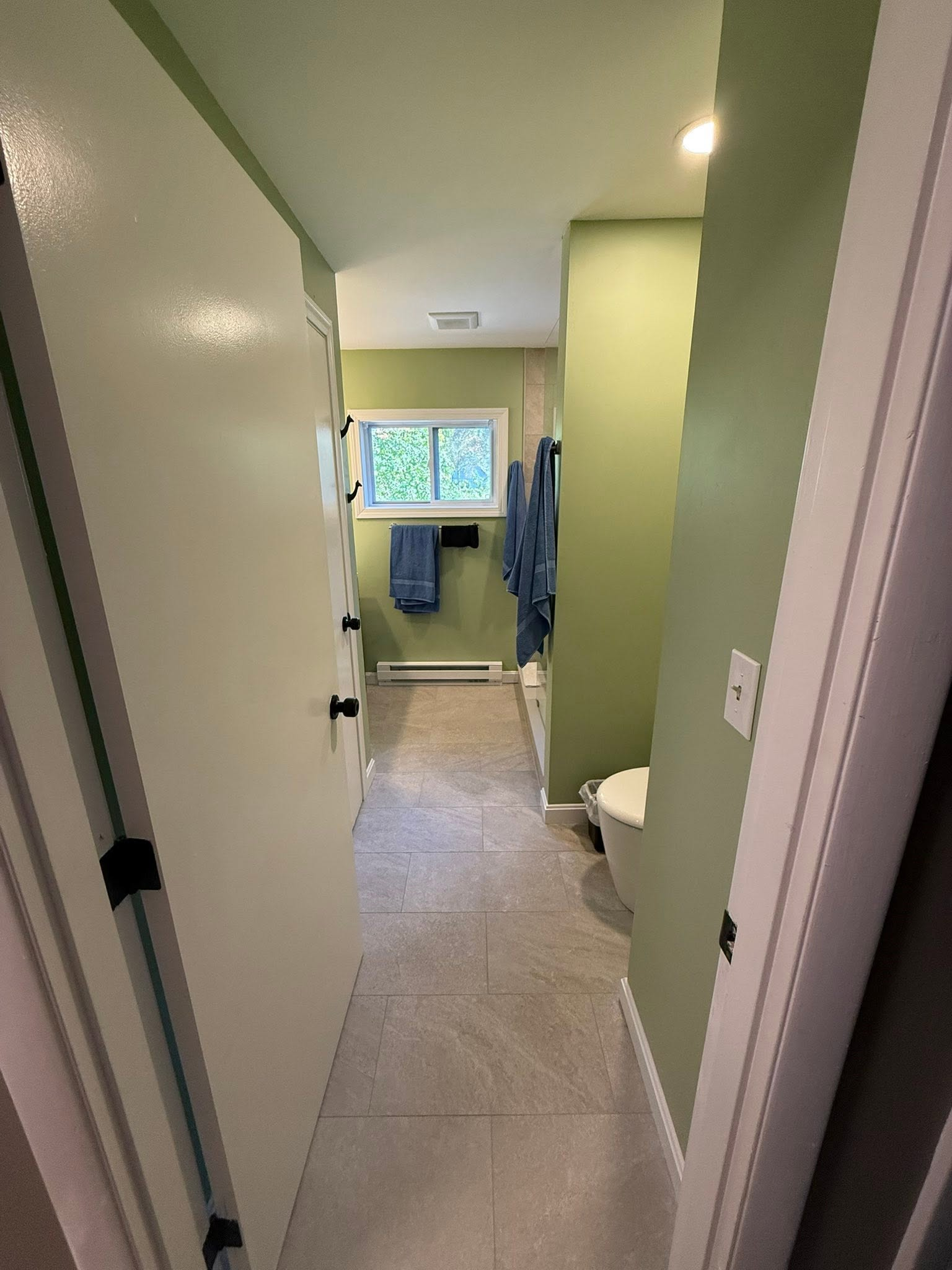 Narrow hallway leading to a bathroom with green walls, tiled floor, and a window at the far end