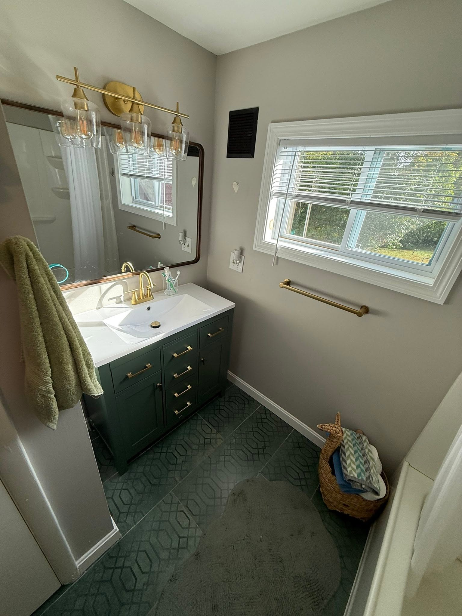Small bathroom with dark green vanity, mirror, white sink, window blinds, and wicker laundry basket.