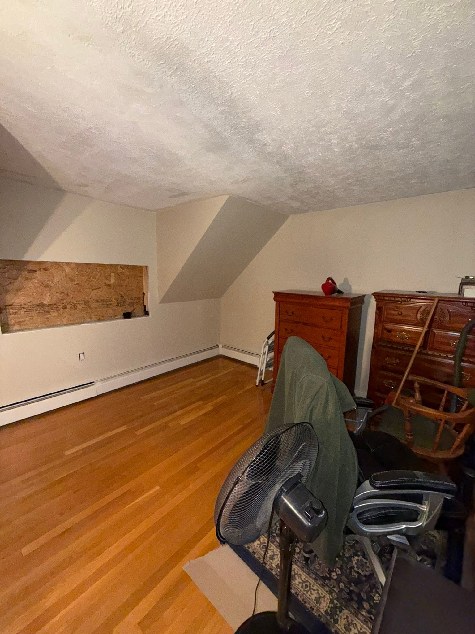 Empty attic room with slanted white ceiling, wooden floor, and furniture stacked on the right.