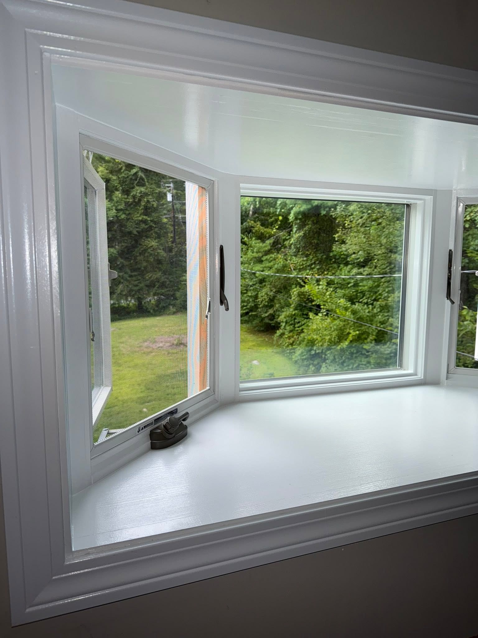 White bay window with open side panels overlooking green trees and lawn