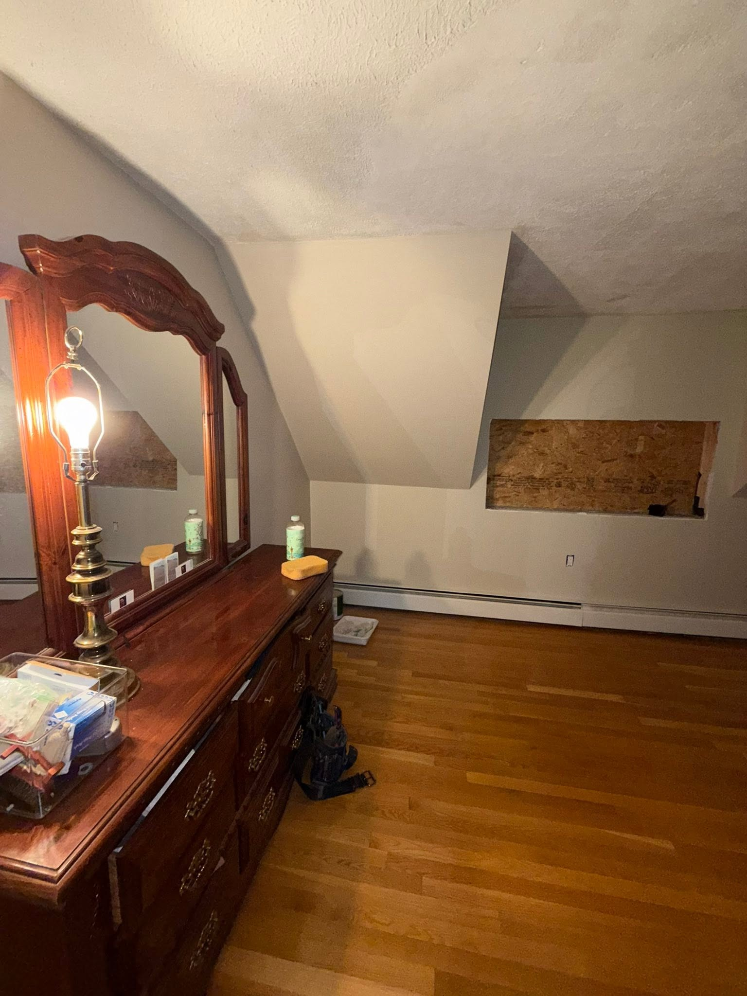 Bedroom with wooden dresser and mirror, hardwood floor, and a window covered by a board.