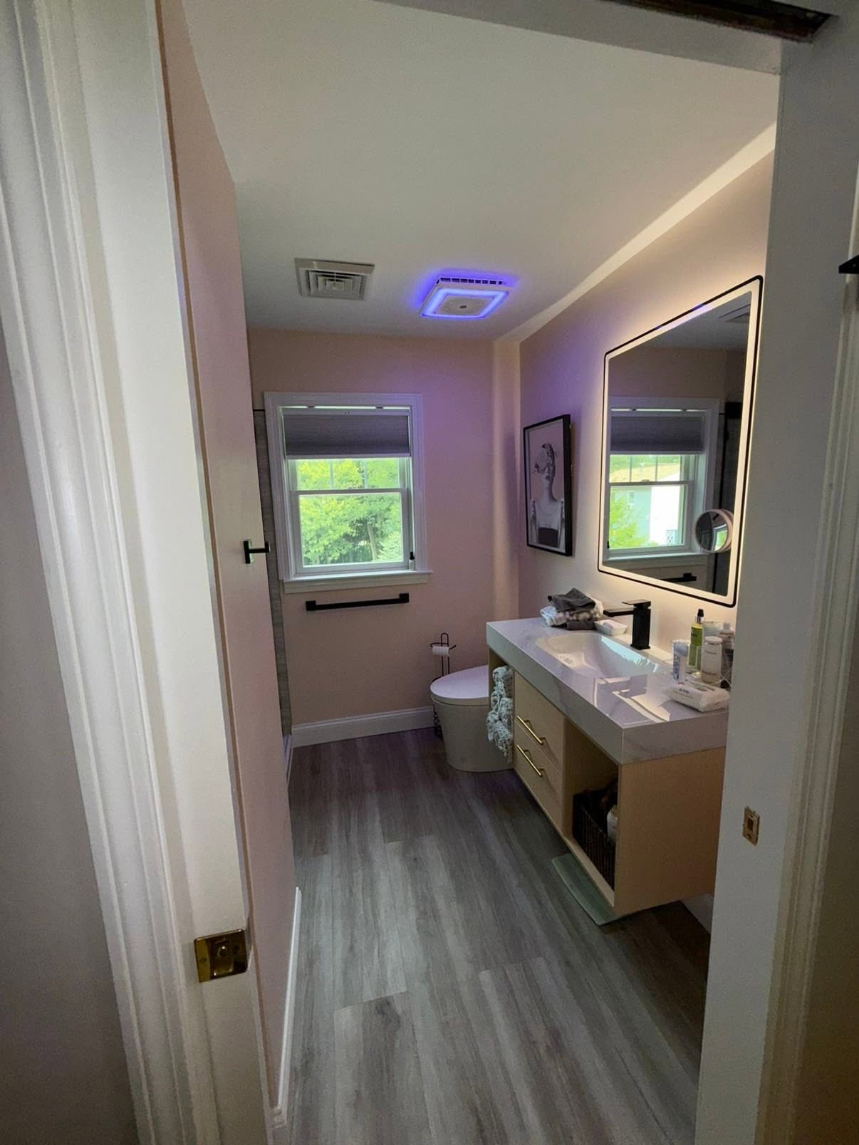 Bathroom with white vanity, large mirror, window, and blue ceiling light in a narrow hallway.