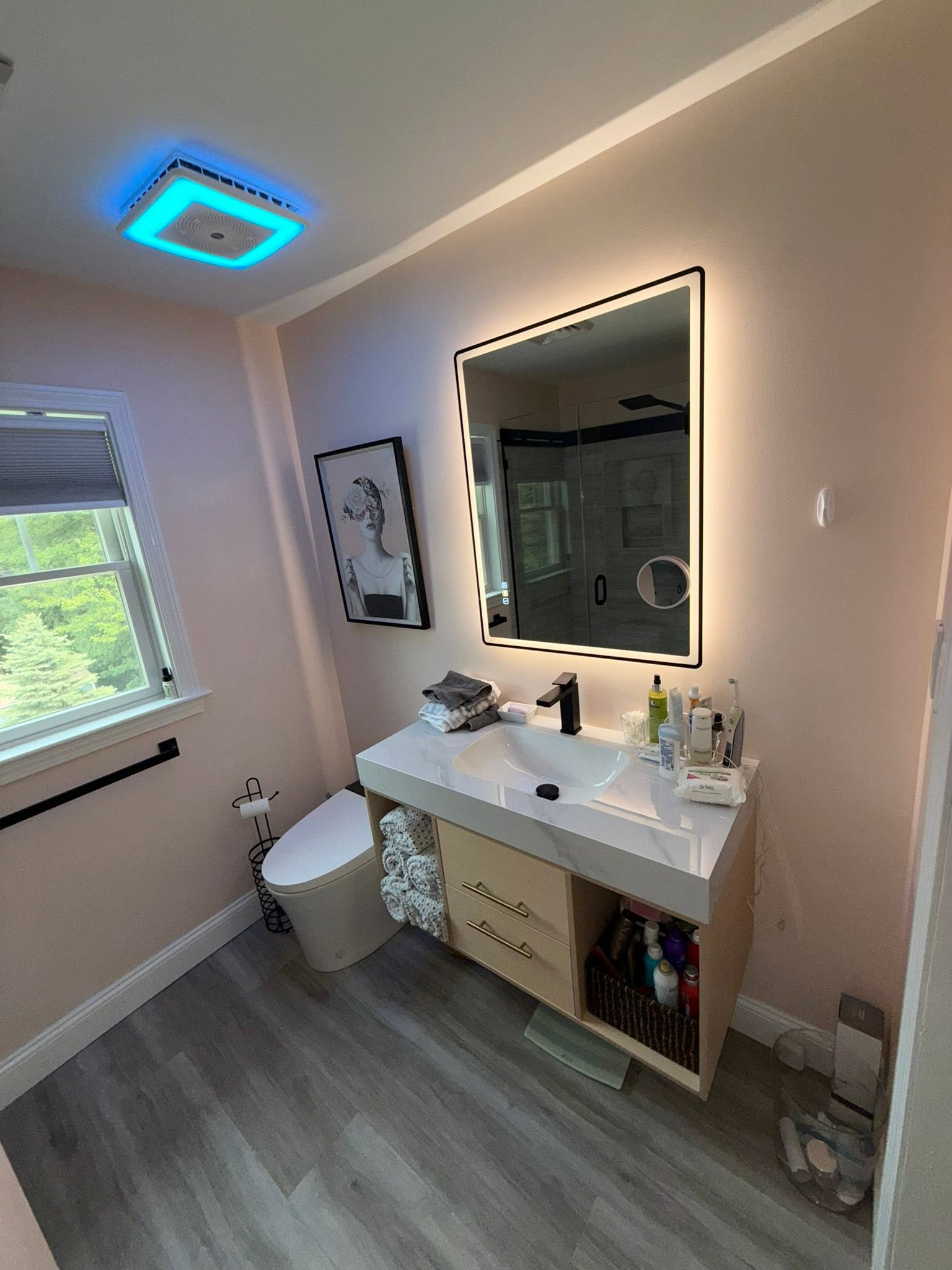 Small bathroom with vanity, mirror, toilet, window, and blue-lit ceiling vent.