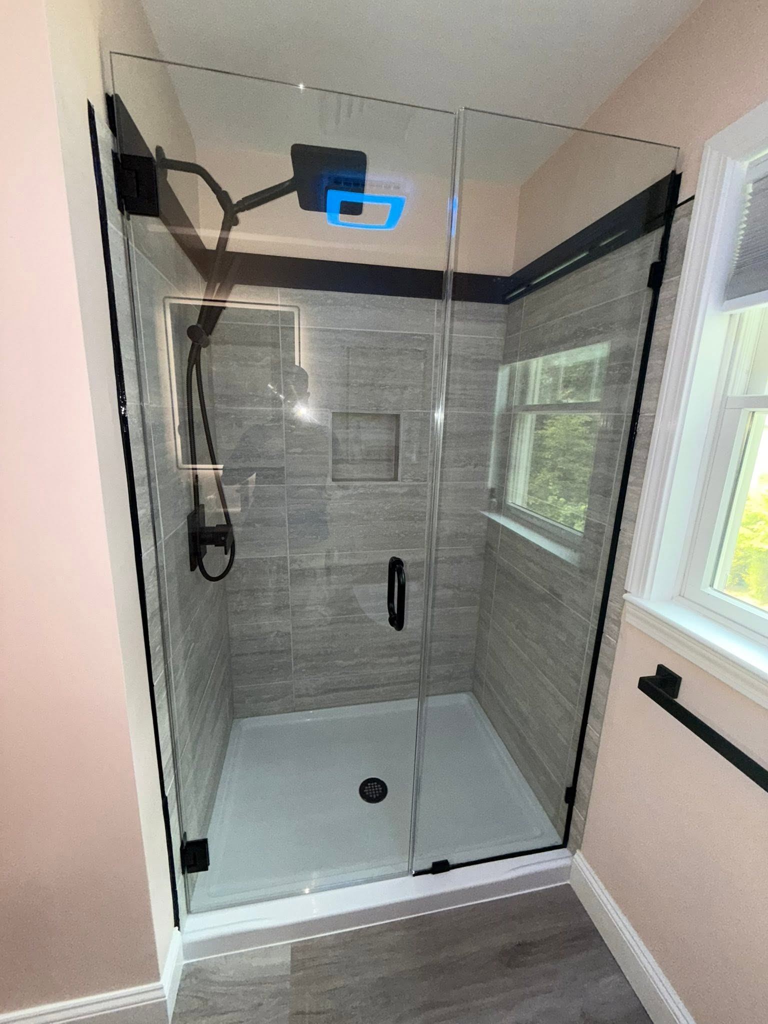 Glass walk-in shower with gray tile walls, white base, black fixtures, and a small window.