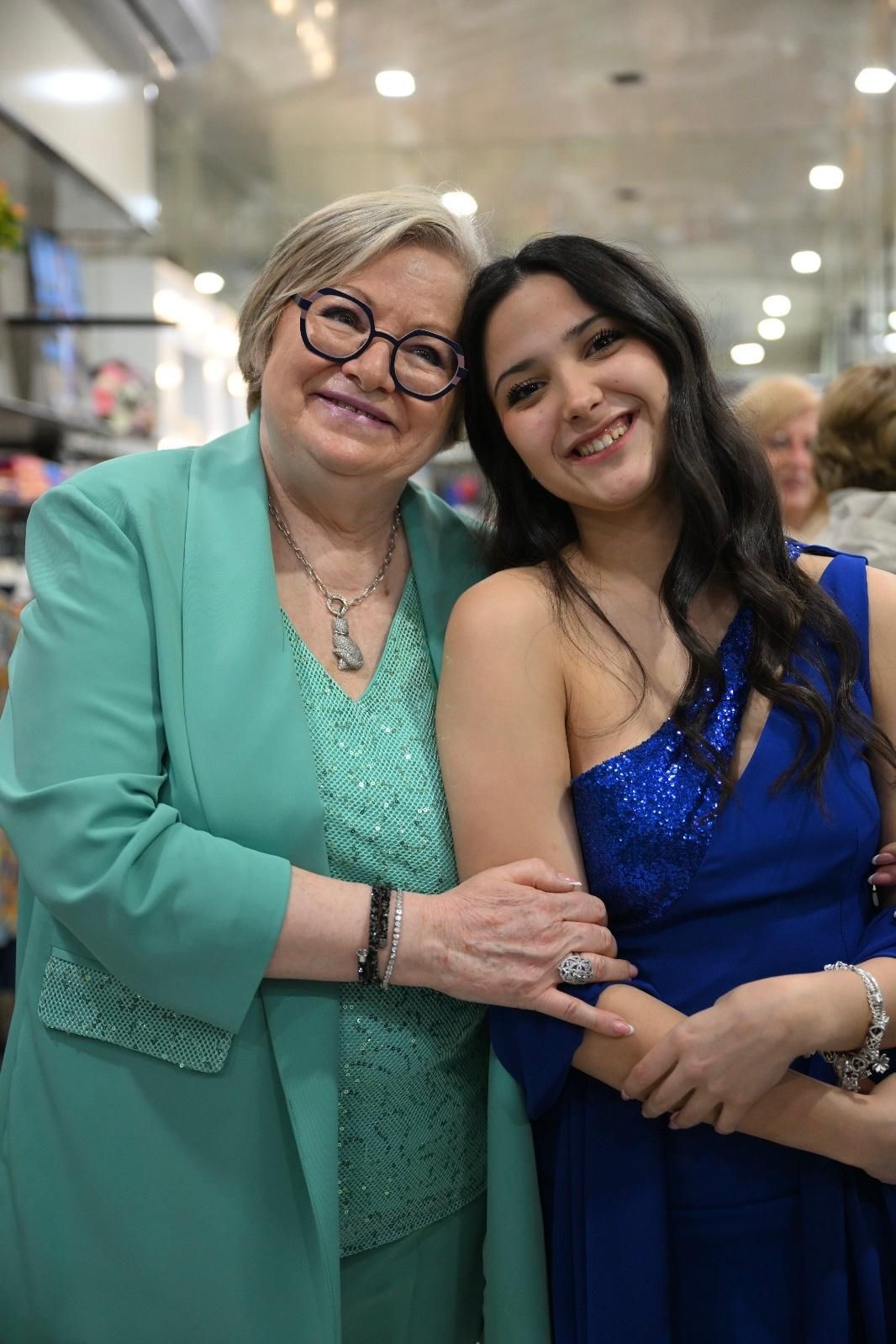 Una donna in abito verde acqua e una giovane donna in abito blu sorridono, a braccetto.