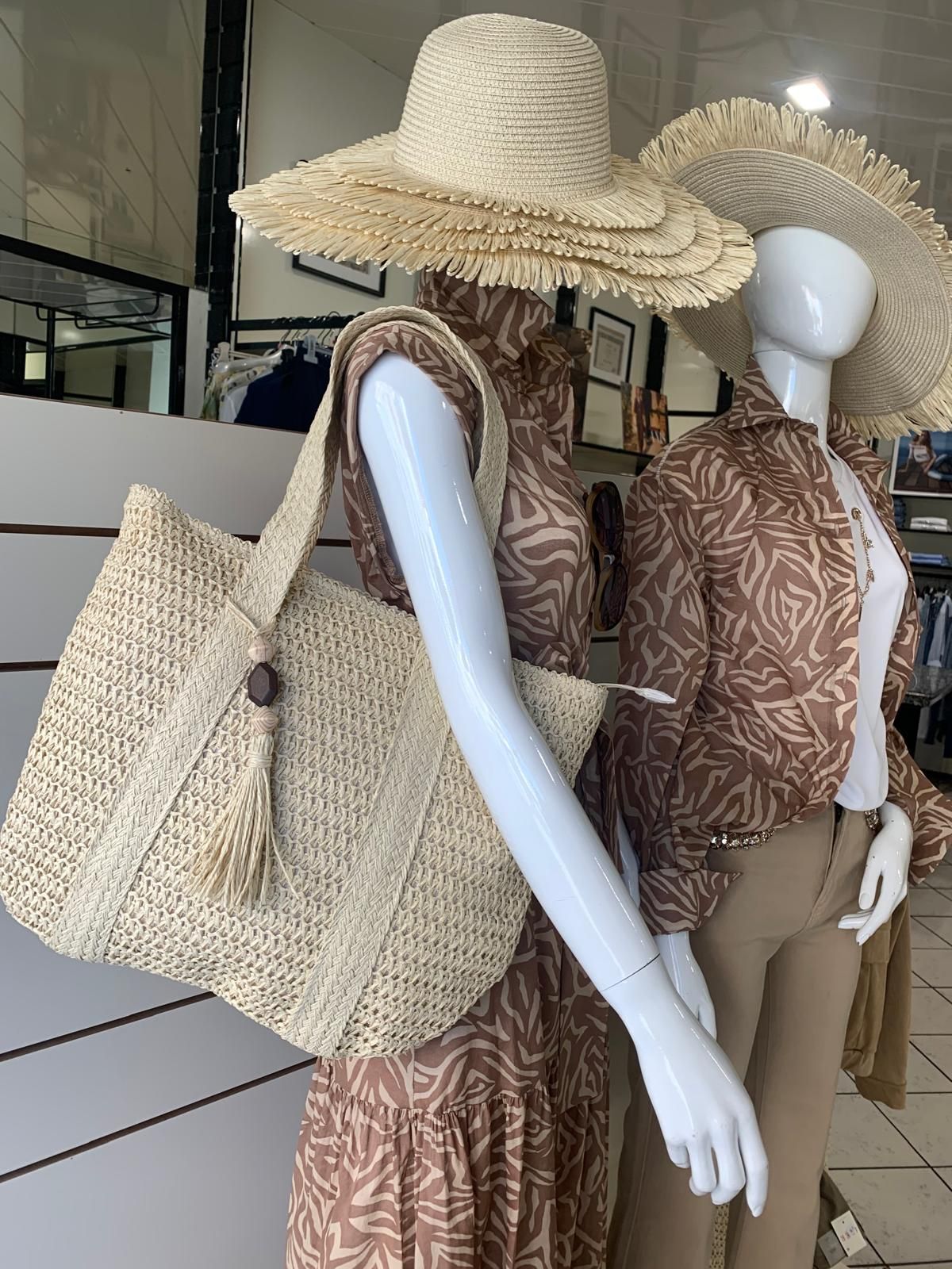 Vetrina di un negozio di abbigliamento con manichini che indossano capispalla e pantaloni chiari. Davanti c'è un piccolo albero di Natale.