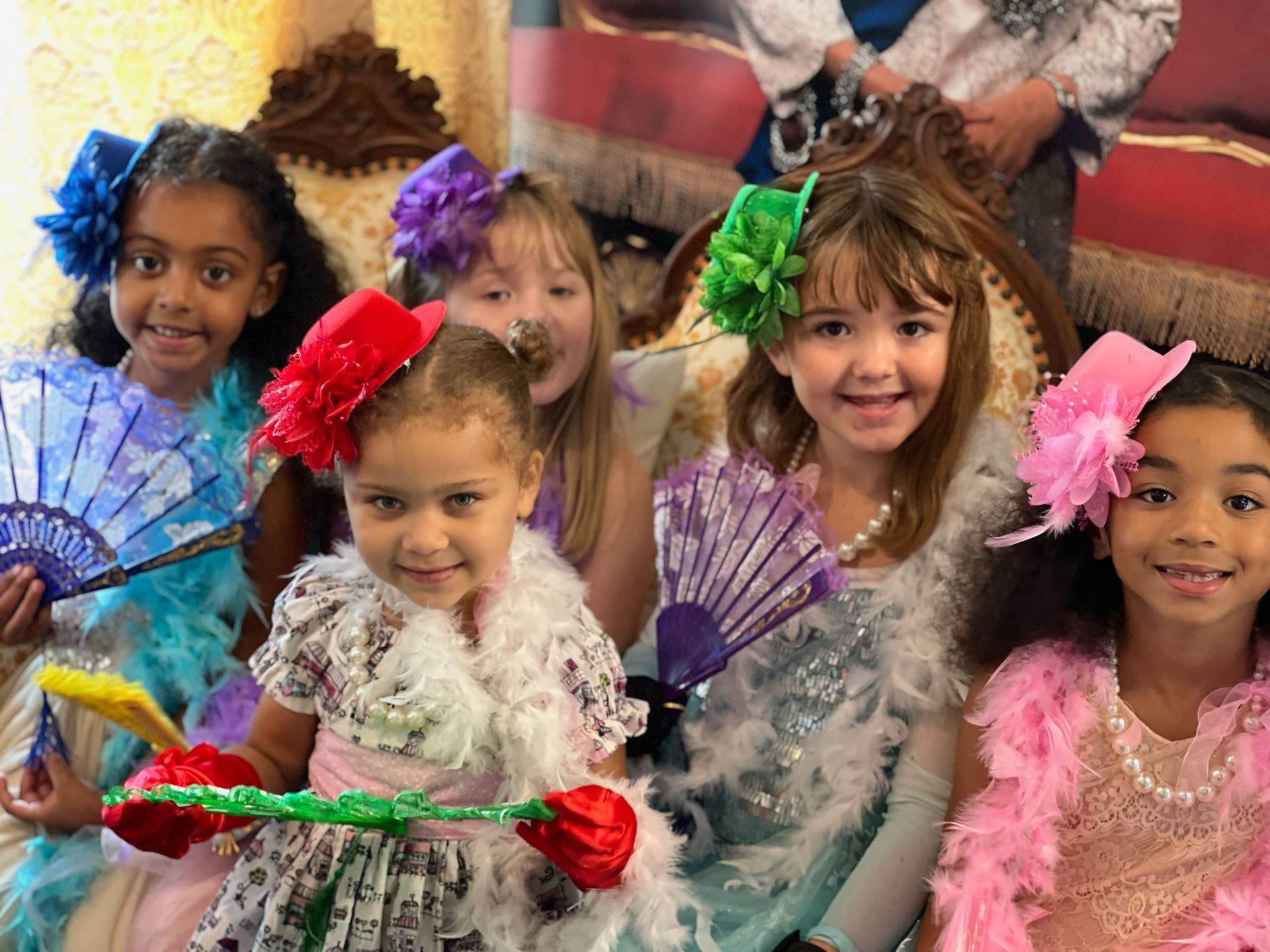 A group of little girls are sitting on a couch holding fans.