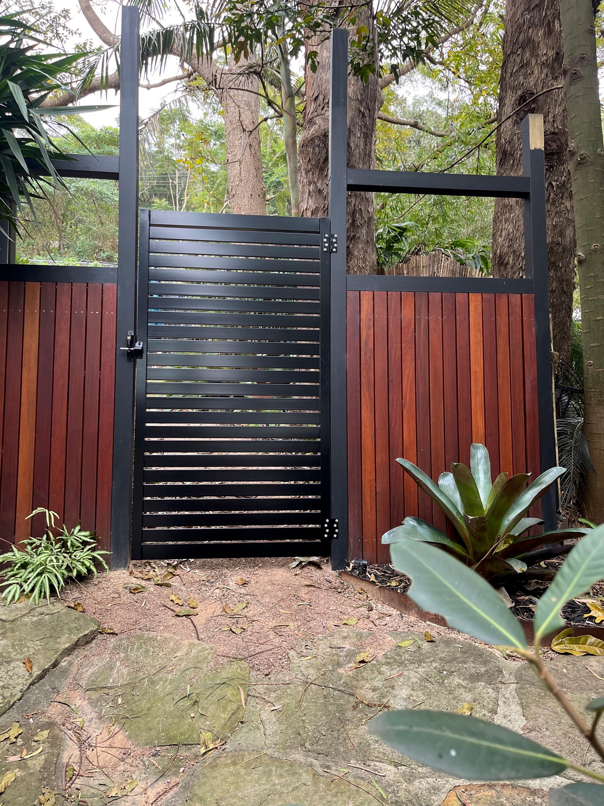 A Wooden Fence With a Black Gate Surrounded by Trees — Febos Welding and Engineering In Corrimal, NSW