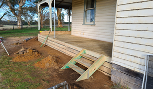 Wooden porch under construction with steps, near a white house with a window, in an outdoor setting.