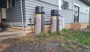 Two water heaters and two propane tanks against a gray house exterior, set in grassy yard.