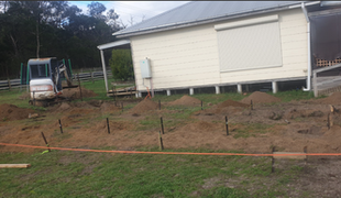 Construction site with mini excavator digging, house on stilts, marked ground for foundation.