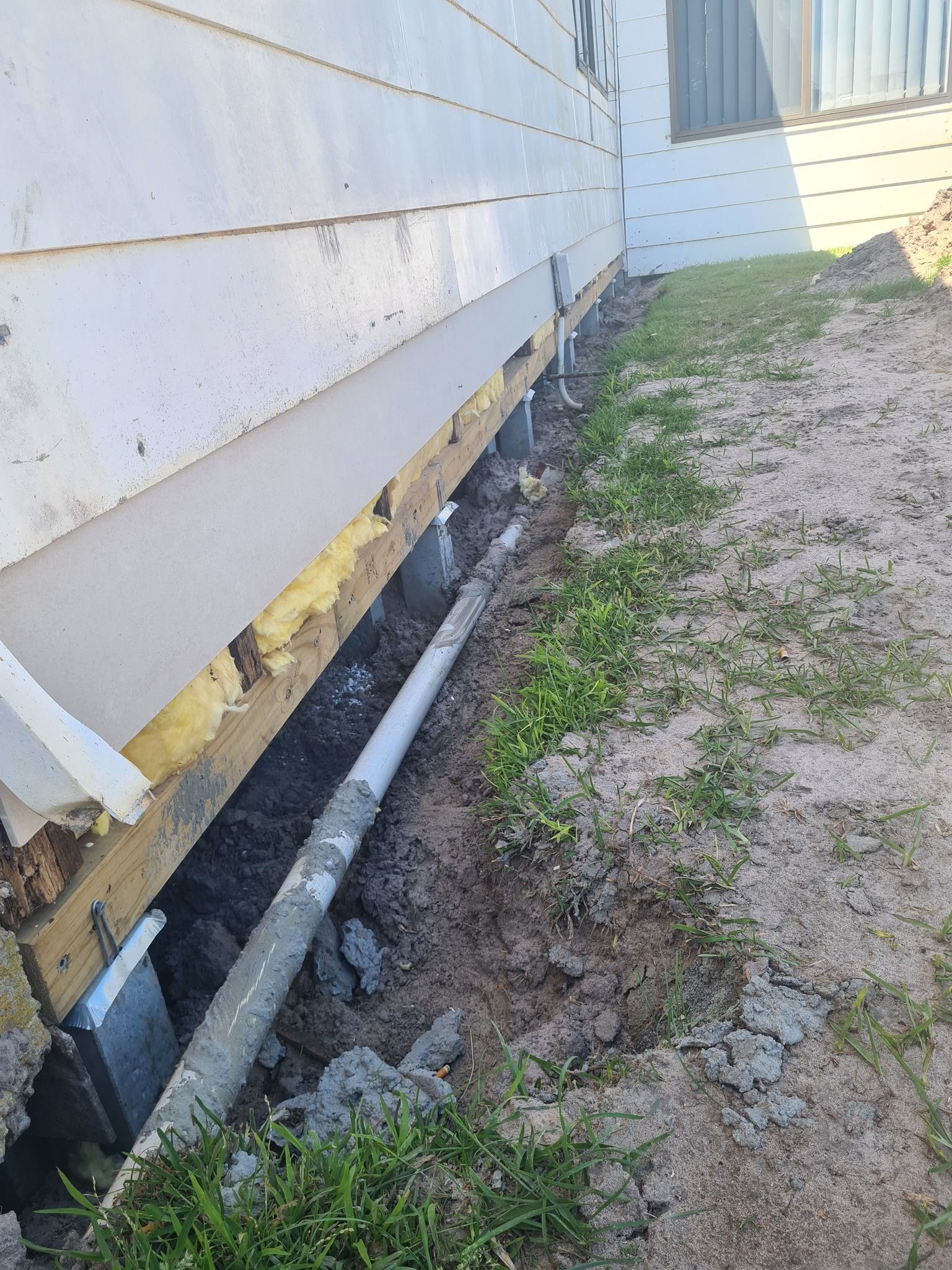 Exterior house wall with exposed foundation, trench, piping, and landscaping.