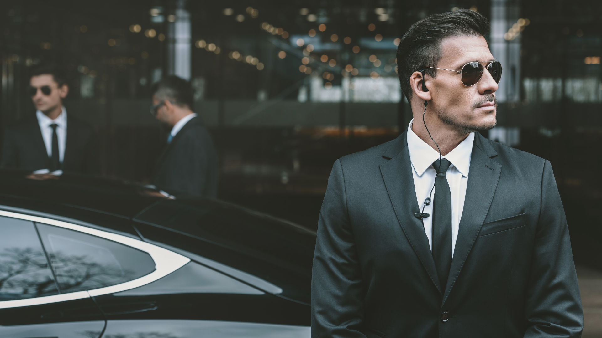 Suited man in sunglasses stands beside a black car outside a modern building.