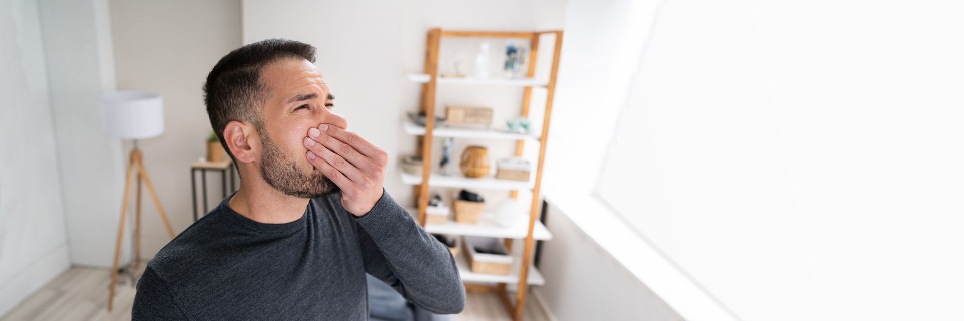 Man sitting in a bright room, covering his mouth as if yawning, with shelves and a lamp behind him