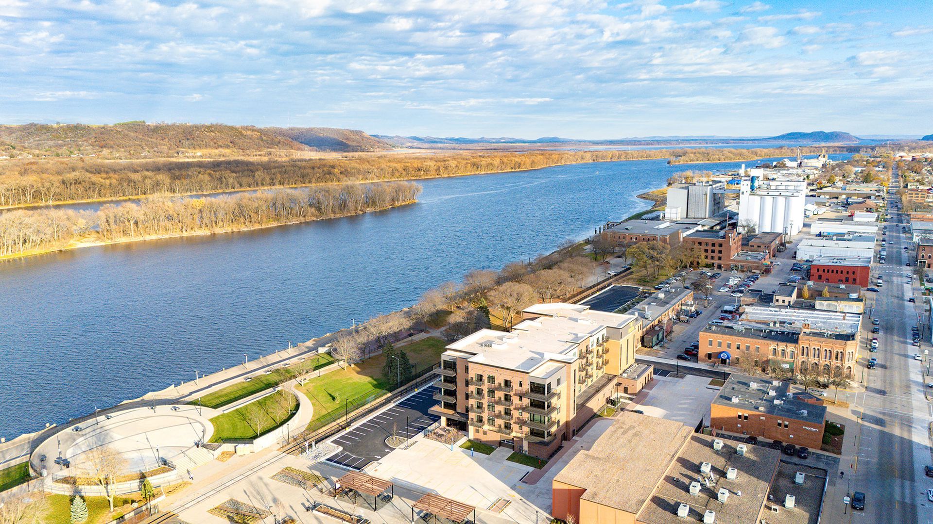Aerial view of a city on a river, with buildings, a park, and a wide blue waterway.