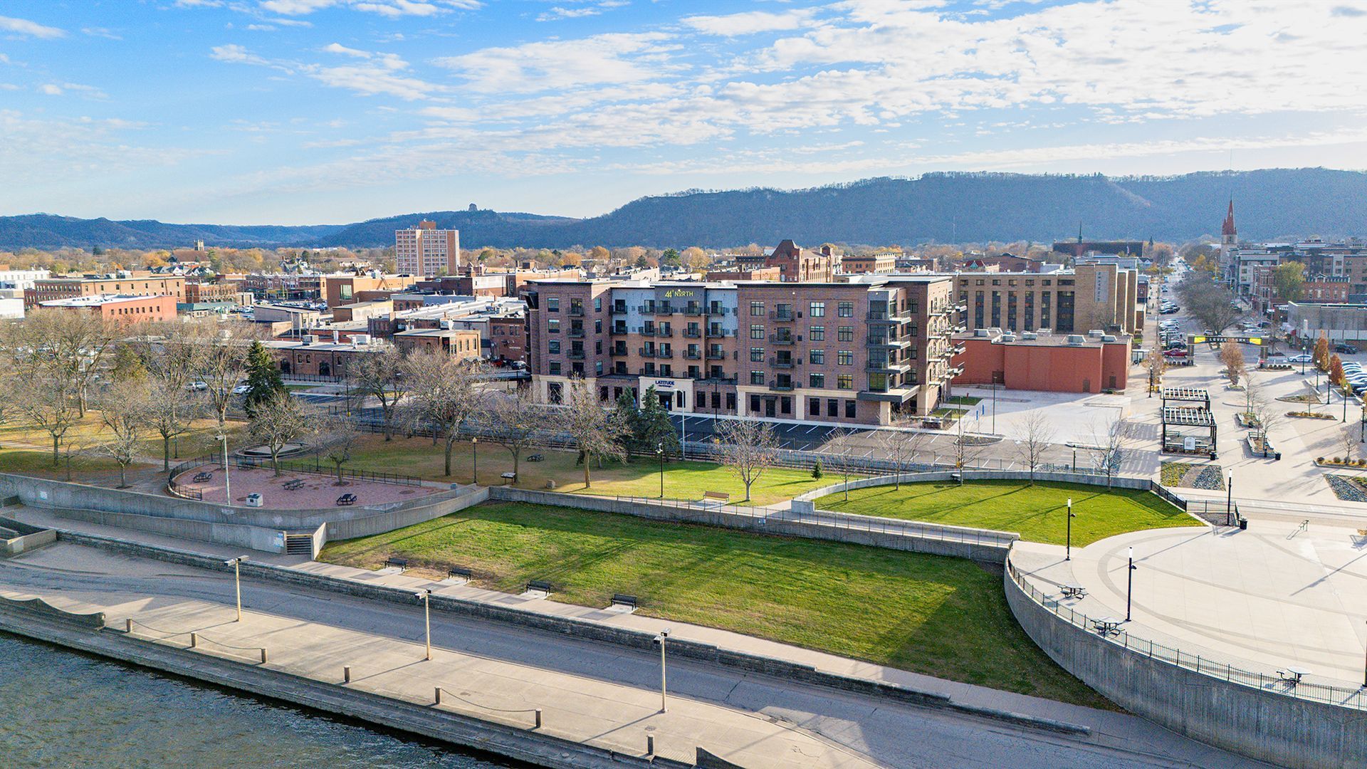 Cityscape with buildings, green space, and a waterfront promenade under a blue sky.