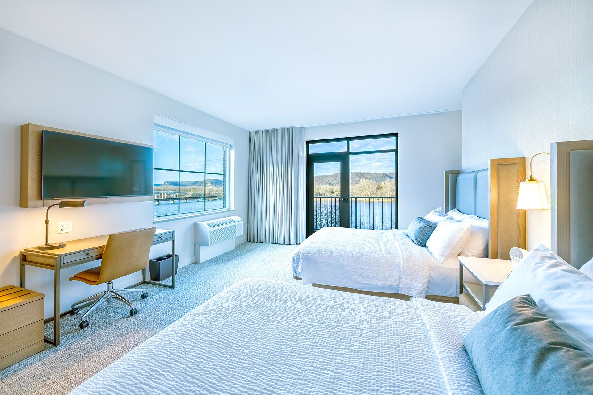 Hotel room with two beds, desk, TV, and window with a view of water and hills.
