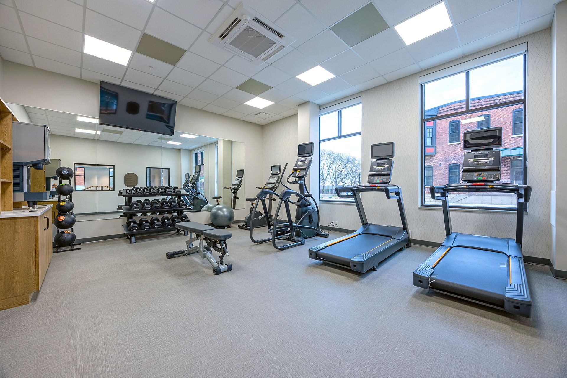 Gym with treadmills, weights, and elliptical machine. Large window with outside view. Gray floor, white ceiling.